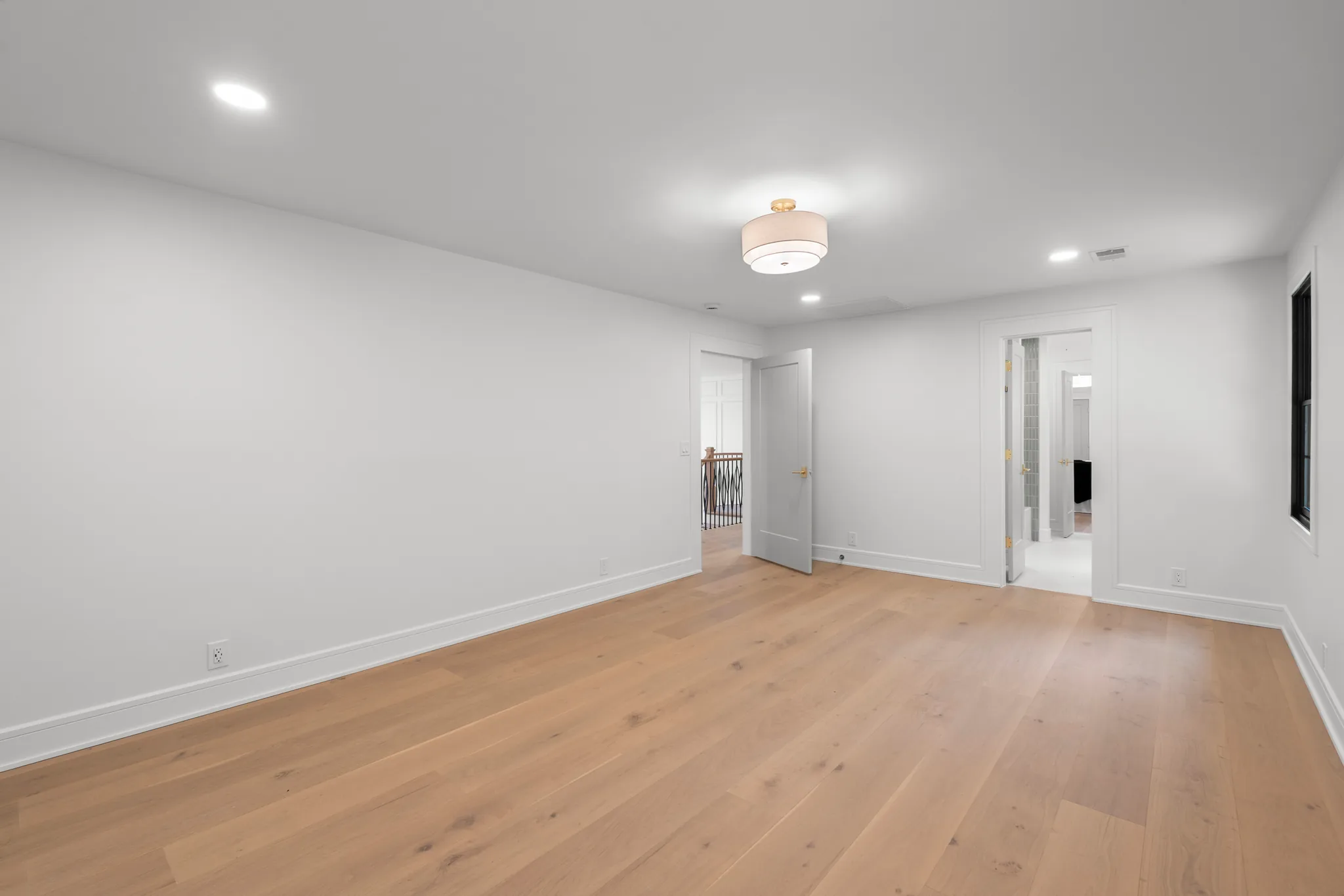 Empty finished bedroom with white walls, wide-plank light oak flooring, drum ceiling light, and open doors to a hallway and tiled bathroom