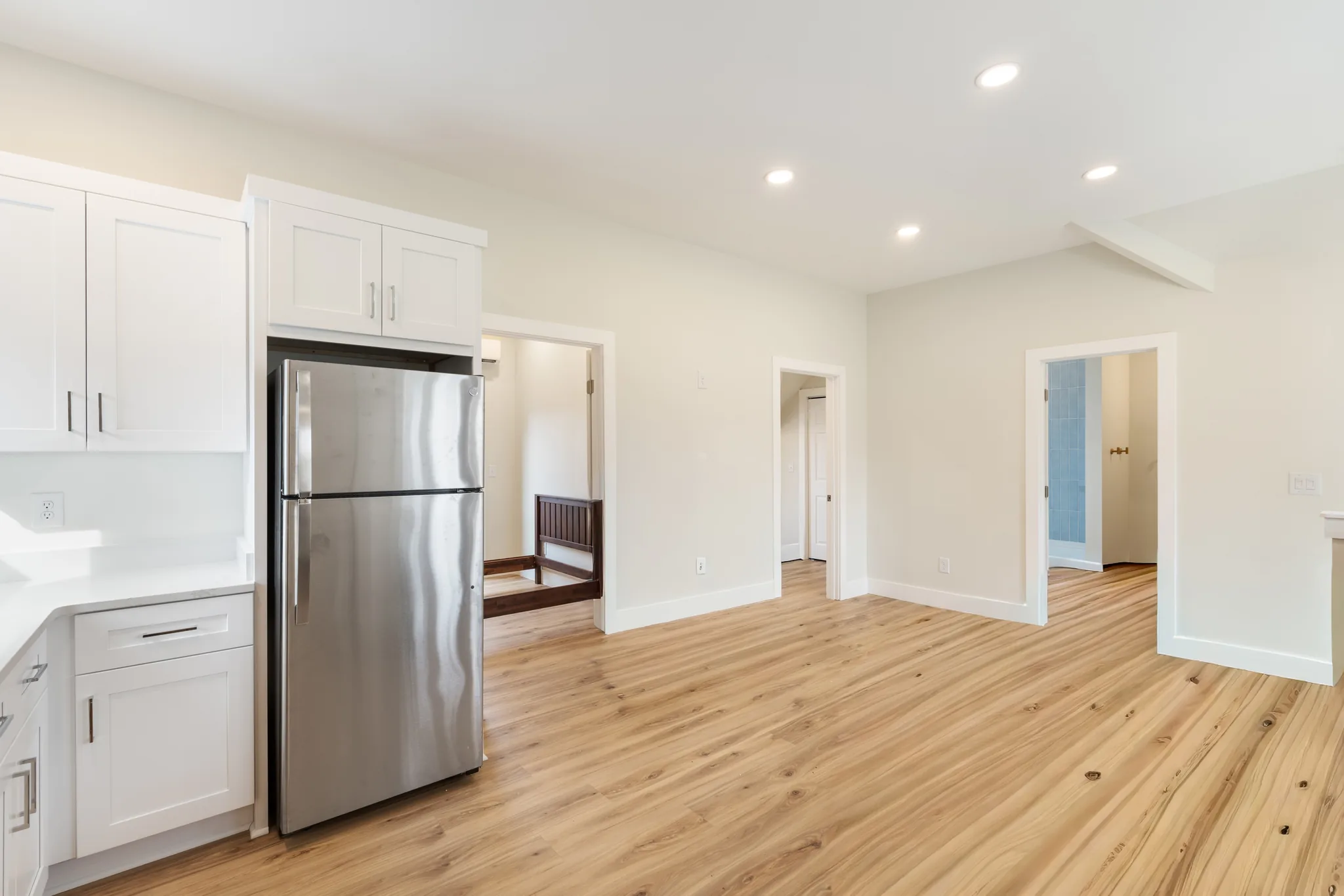 Vacant living area with light wood plank flooring, white shaker cabinets, stainless refrigerator, recessed ceiling lights and open doorways to adjoining rooms