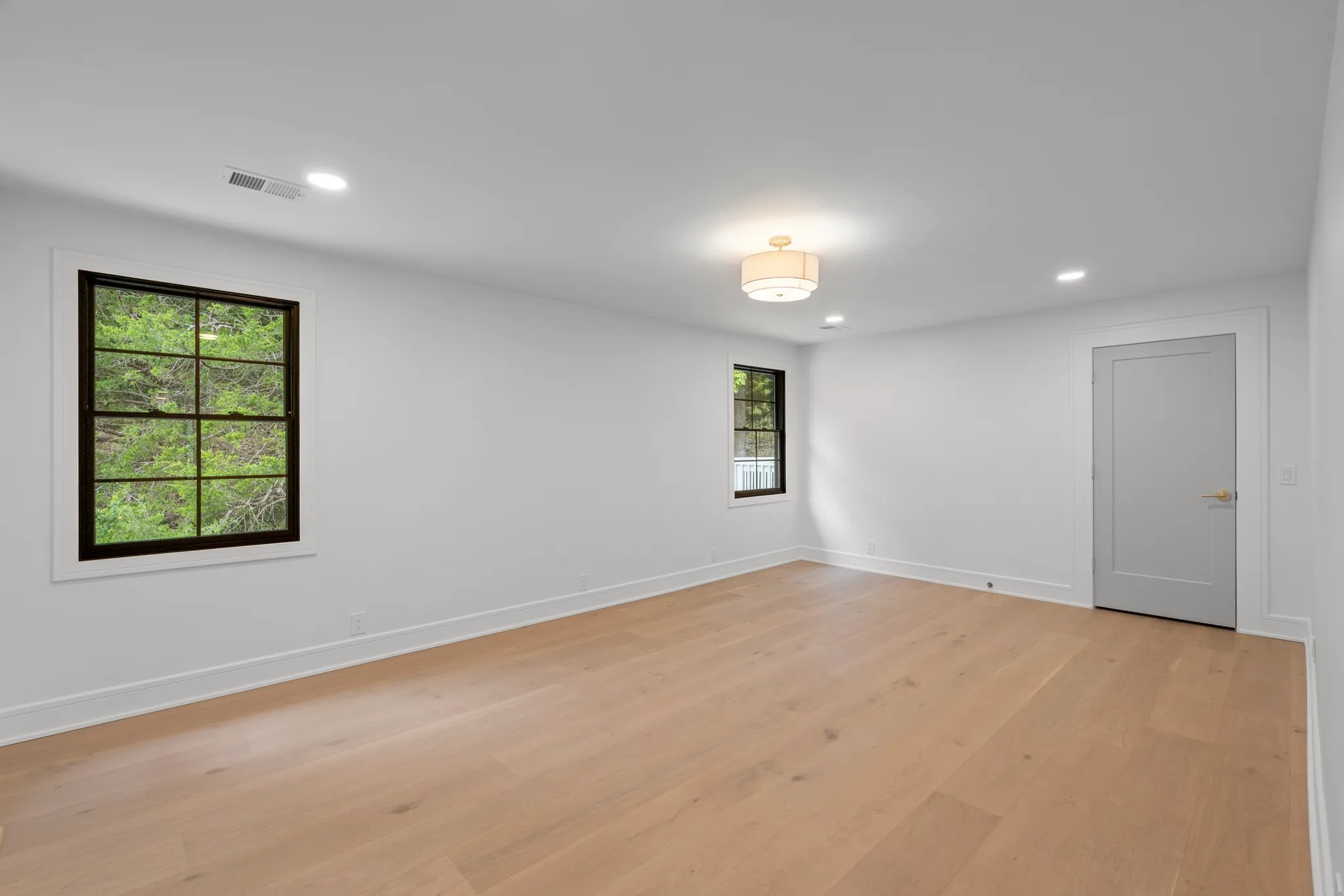 Empty room with white walls, light oak plank flooring, two black grid windows, a gray paneled door and a drum ceiling light