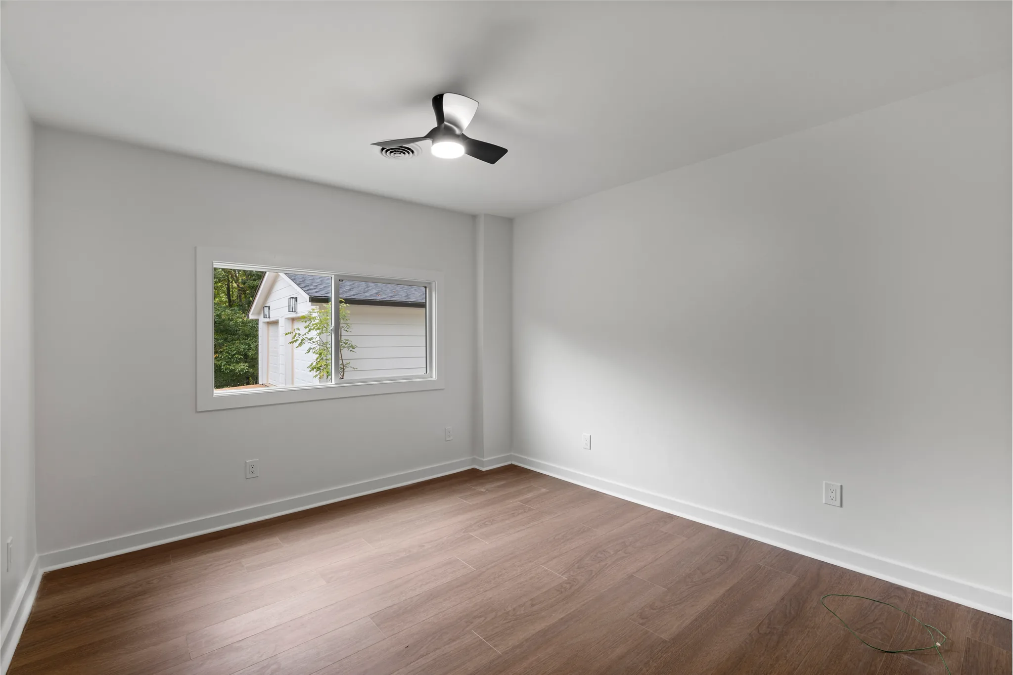 Empty room with light gray walls, wood-look plank flooring, white baseboards and a black and white ceiling fan beside a sliding window