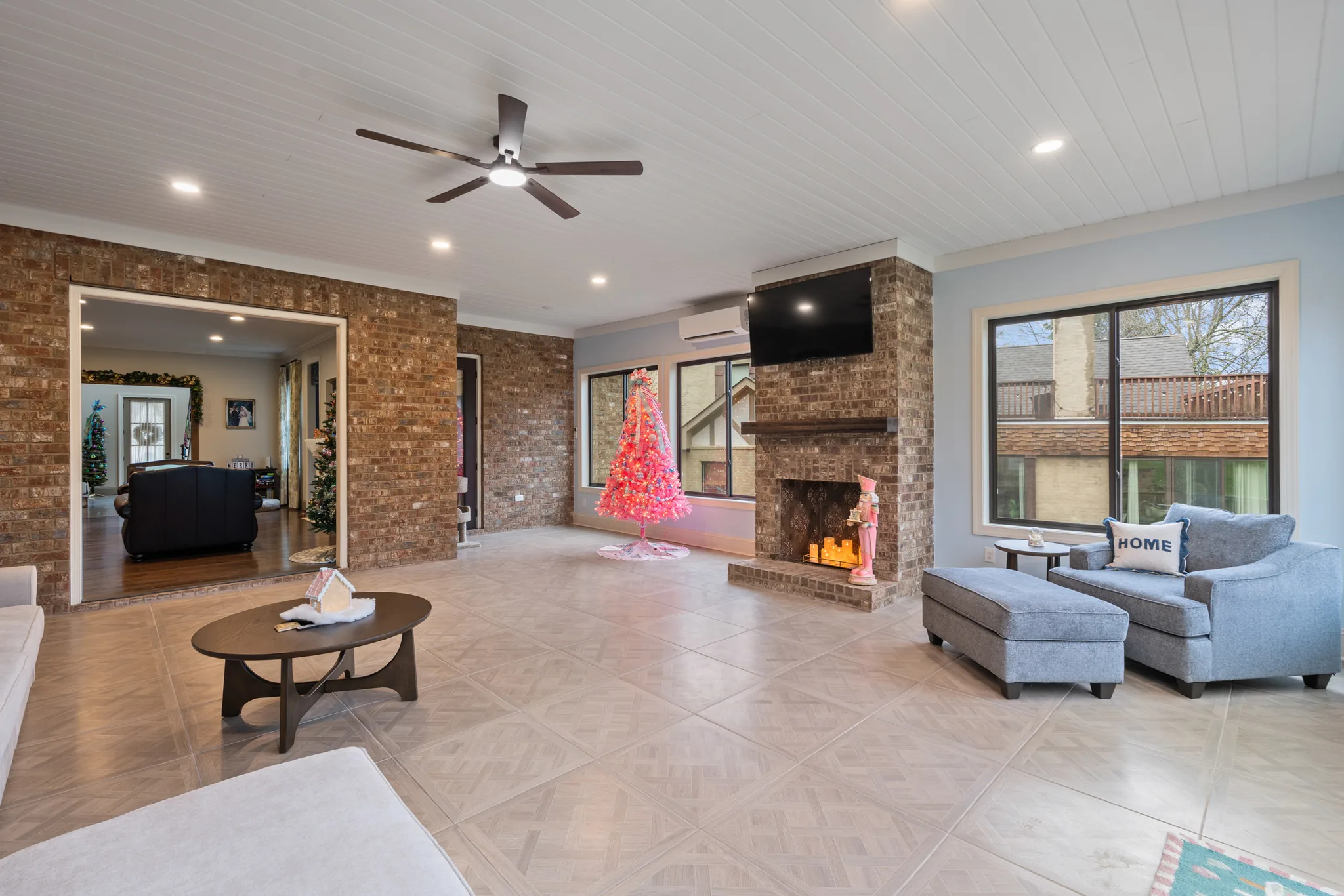 Enclosed porch with a brick fireplace and mantel, white plank ceiling with fan, large tile floor and a wide opening into the living room