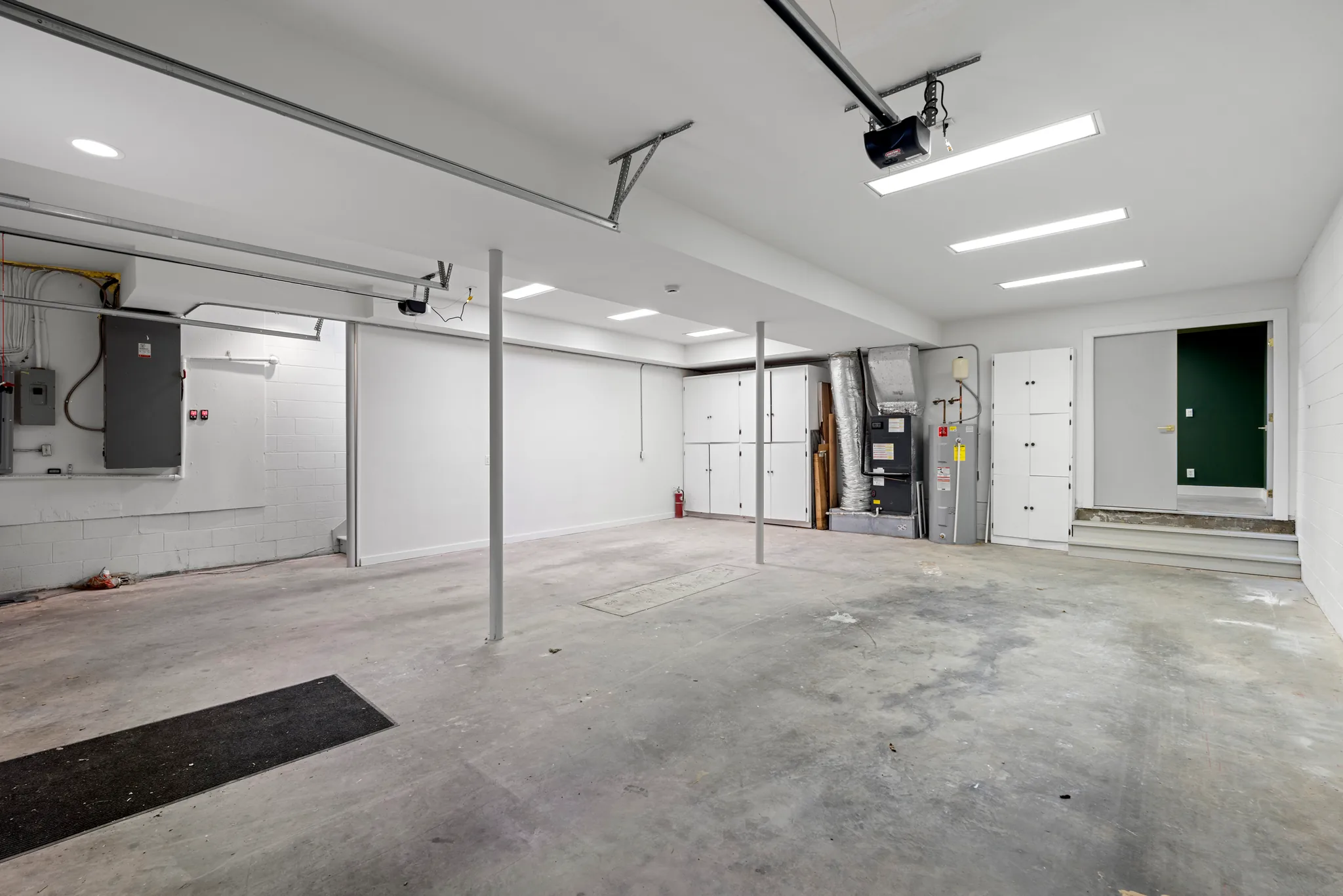 Finished basement-level garage with white painted block walls, steel support posts, concrete floor, white storage cabinets, water heater and green door beyond steps