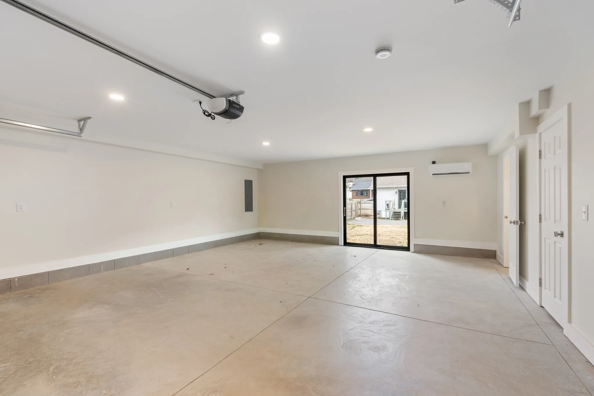 Empty finished garage interior with white walls, recessed ceiling lights, opener rail, sliding glass door to a fenced yard and sealed concrete floor