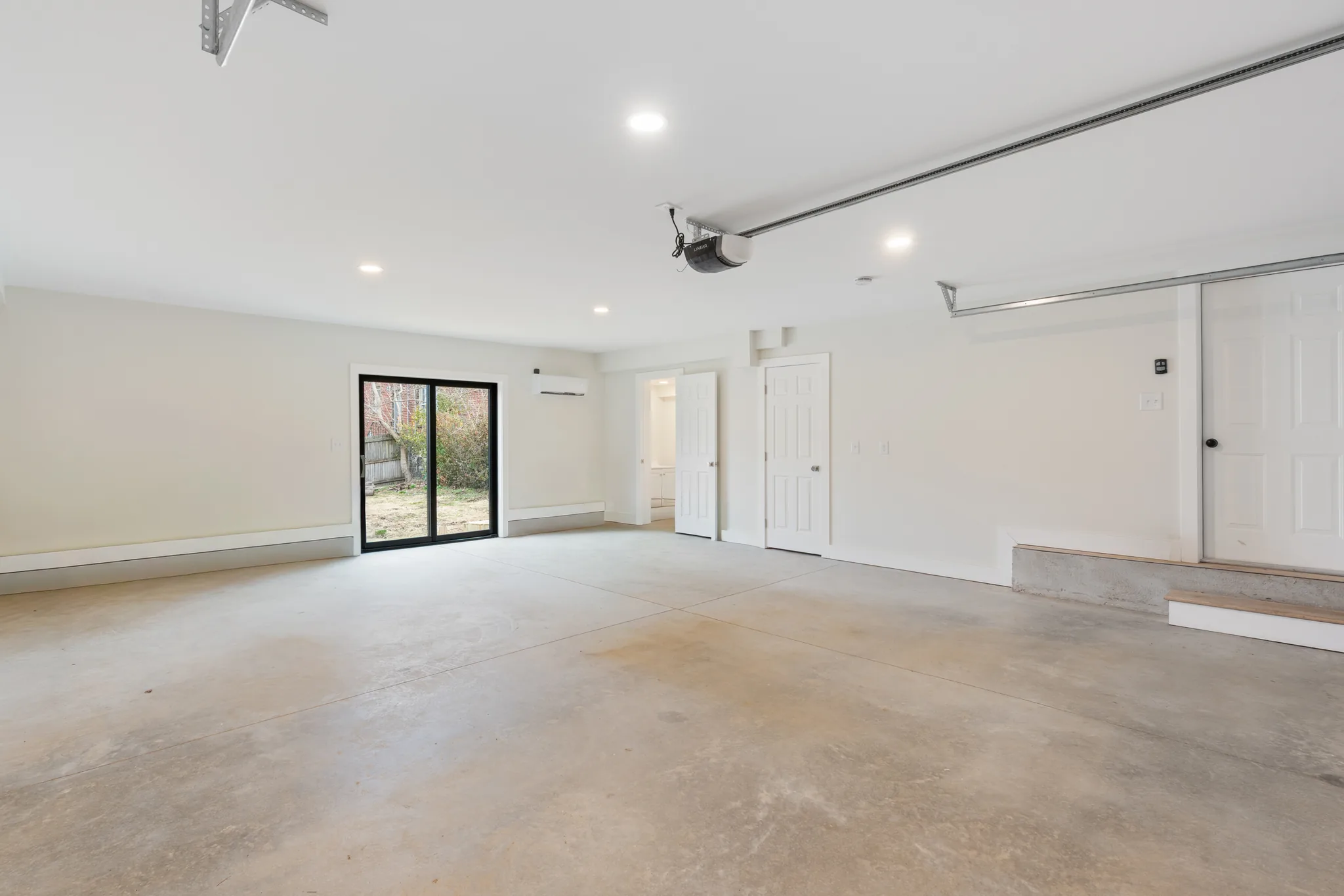 Empty finished garage with white walls, polished concrete floor, sliding glass patio door to the yard and two white six-panel doors