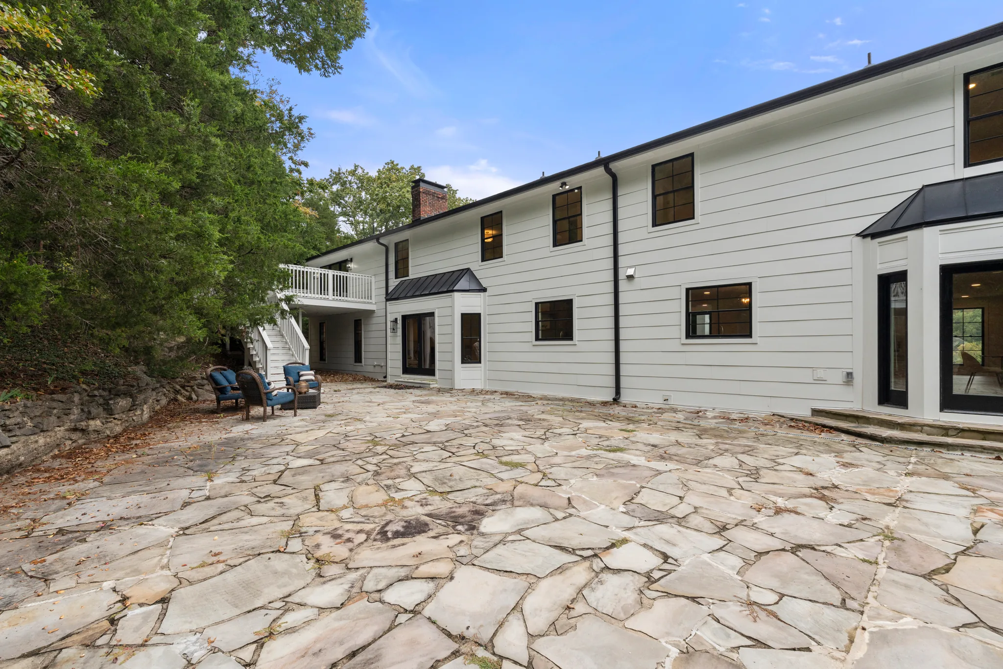 Large irregular flagstone patio behind a two-story white lap-siding home with black windows, a small upper deck and wicker seating near a stone retaining wall