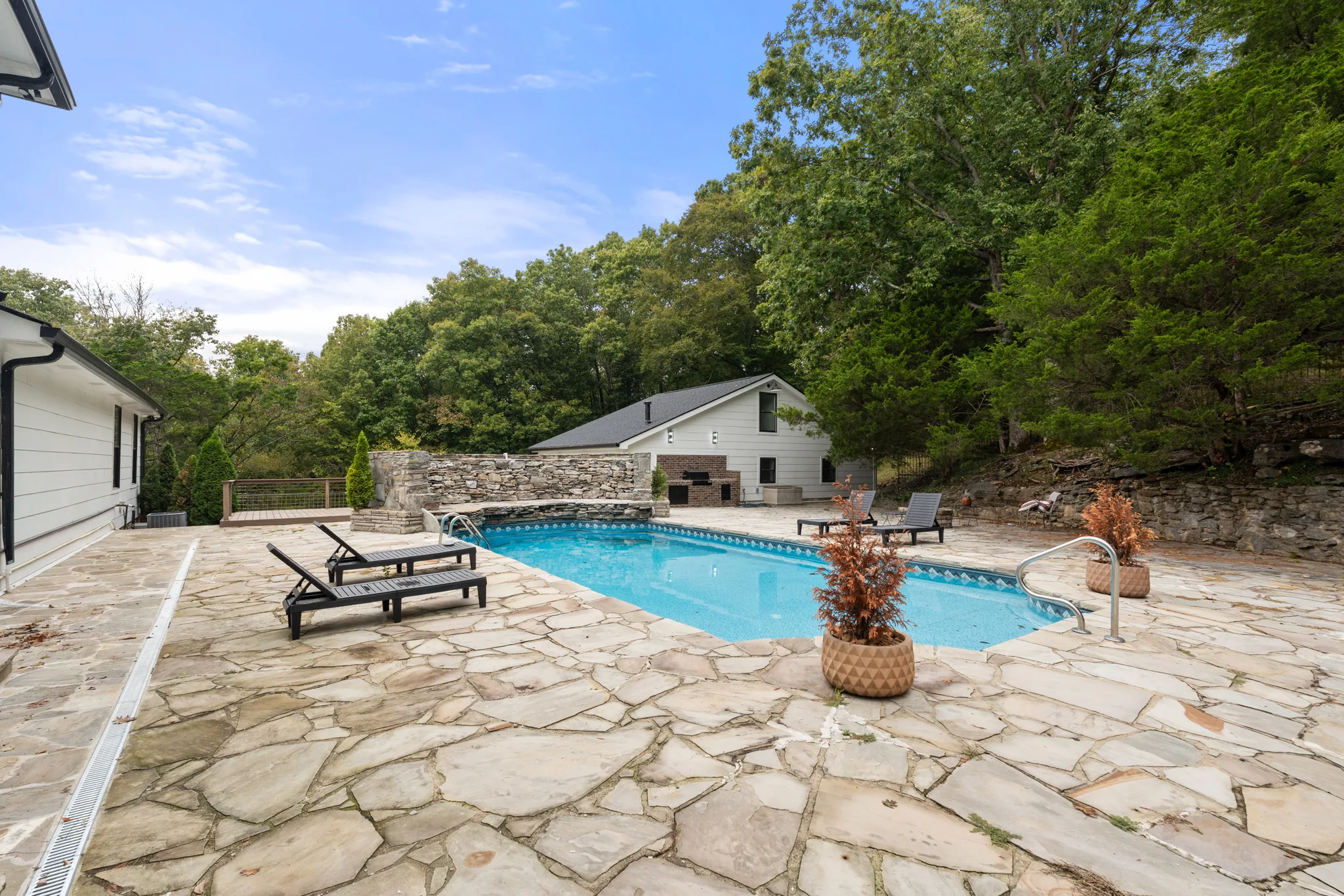 In-ground pool surrounded by a wide flagstone patio with lounge chairs, a stone retaining wall and a white outbuilding among trees