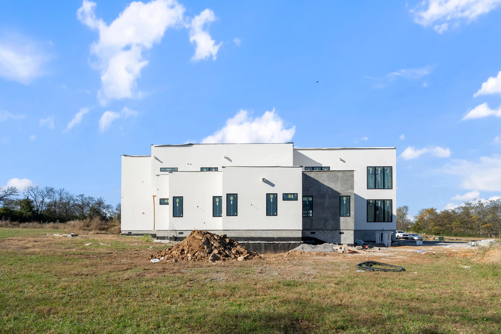 White stucco flat-roof home under construction in an open field, with tall dark-framed windows, a gray accent wall and a dirt pile in front