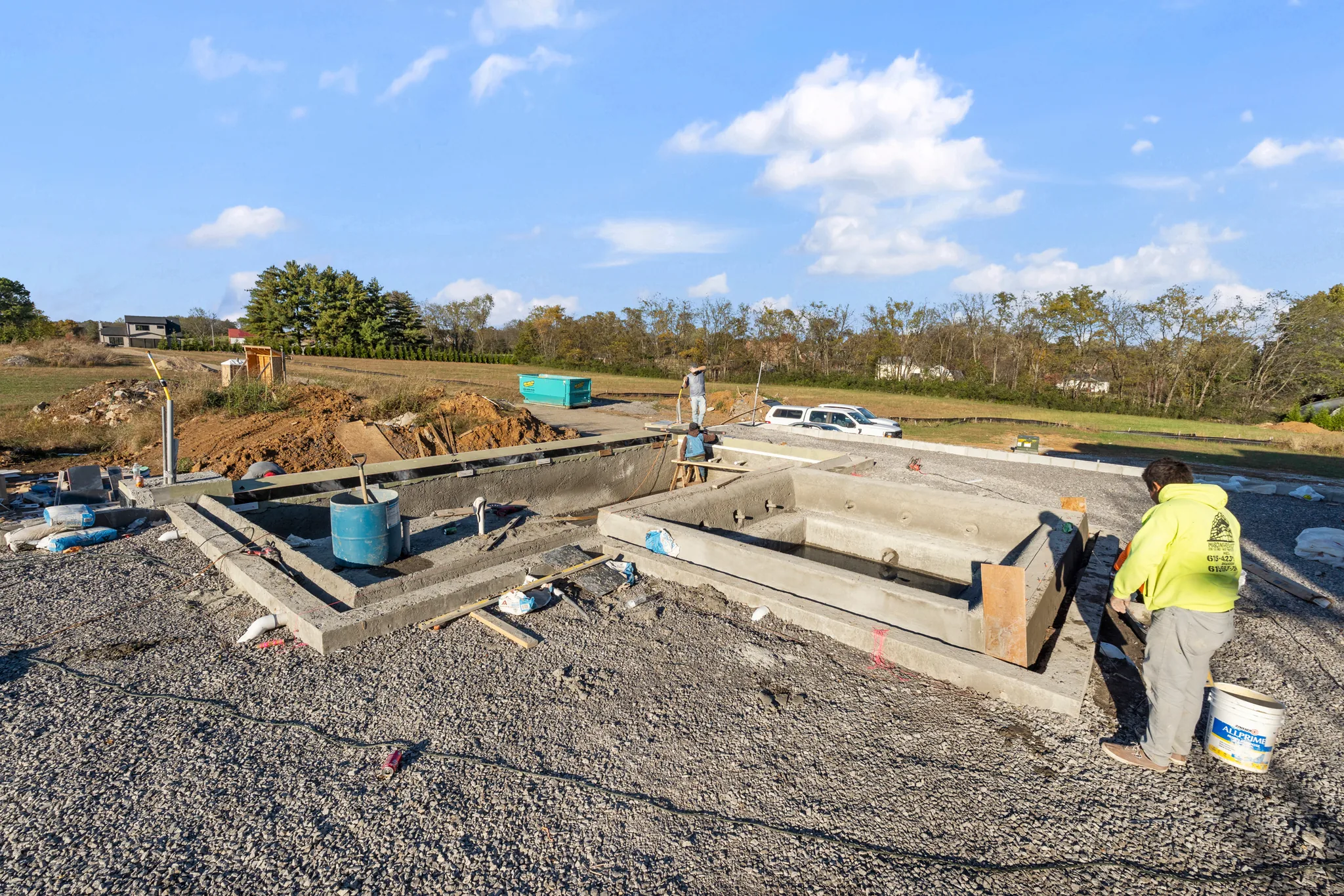 Concrete foundation walls and footings under construction on a gravel pad, with workers in high-visibility clothing and buckets and lumber nearby