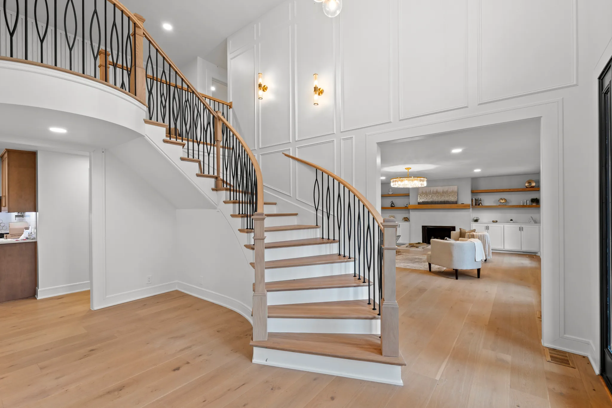Two-story entry with a curved staircase, oak handrail, black metal balusters, white wall paneling and wide-plank oak floors leading to a living room
