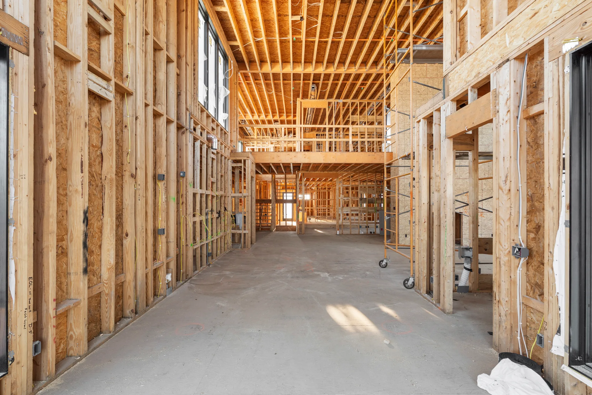 Interior framing under construction showing a wide concrete floor, stud walls with rough wiring, scaffolding and an open second-floor bridge above