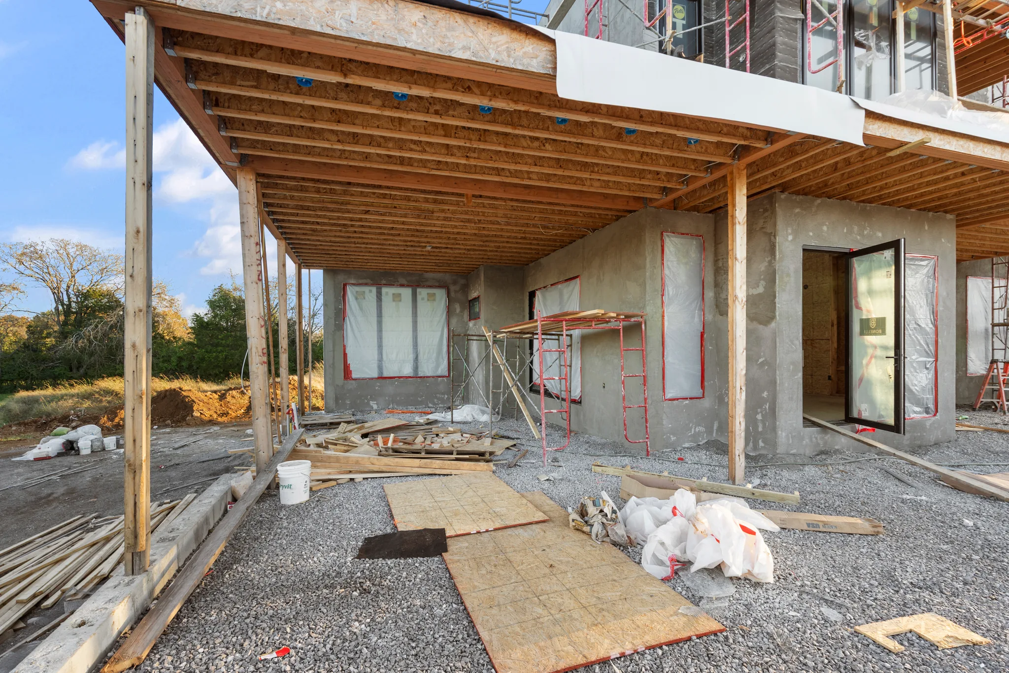 New home under construction with exposed floor joists over a concrete lower level, taped window openings, gravel grade and scaffolding