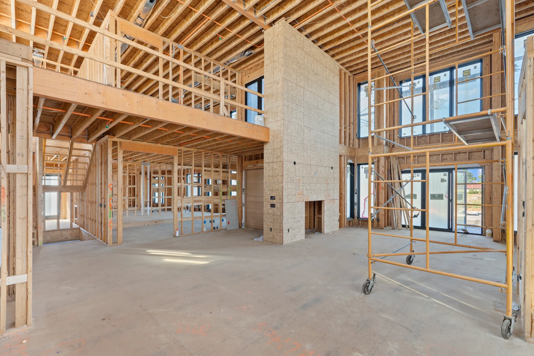 Two-story great room under construction with wood framing, a sheathed fireplace chase, loft walkway above, black windows and rolling scaffold on a concrete slab