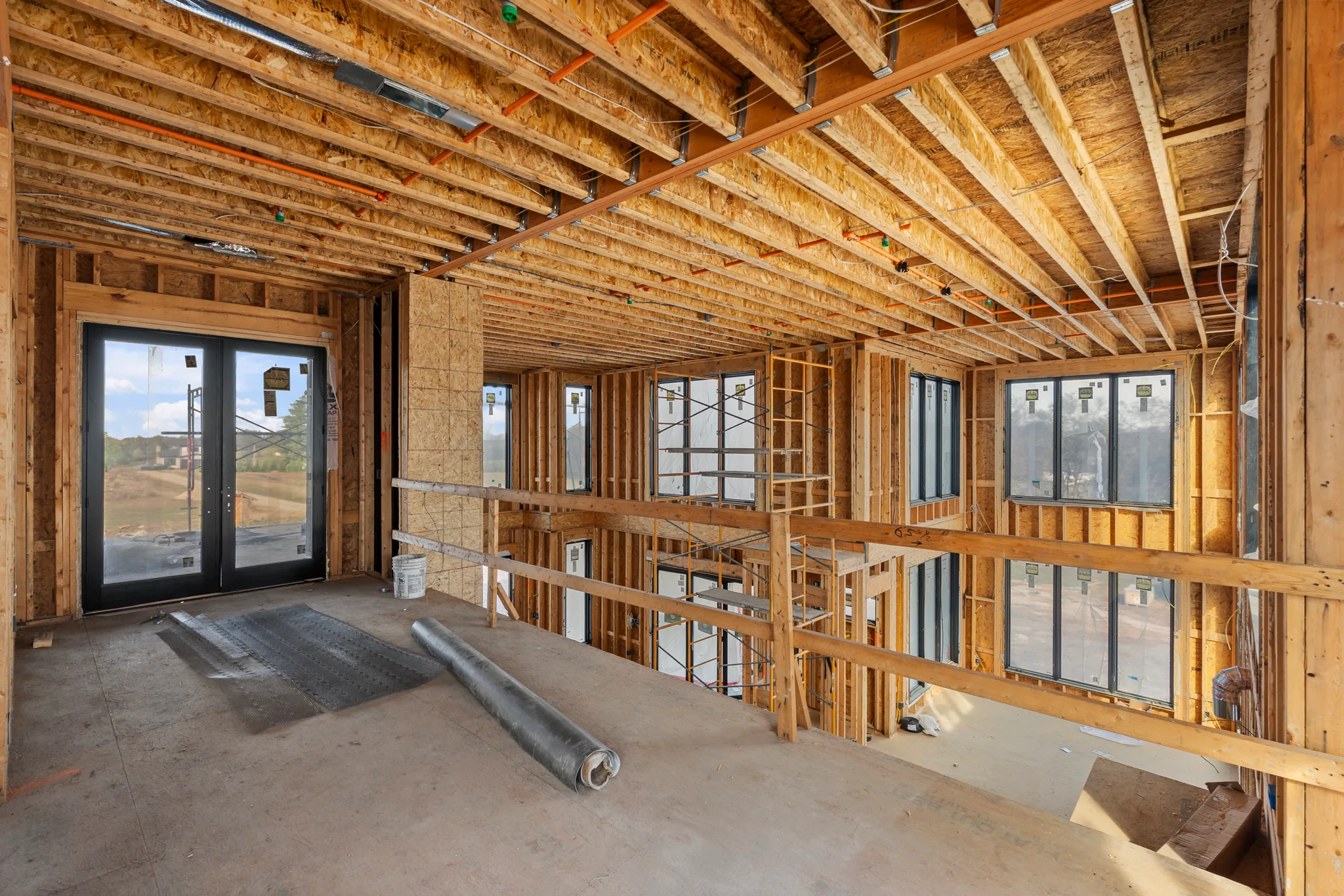 Interior framing under construction with exposed joists, rough wiring, black French patio doors and multiple black window units overlooking a cleared site