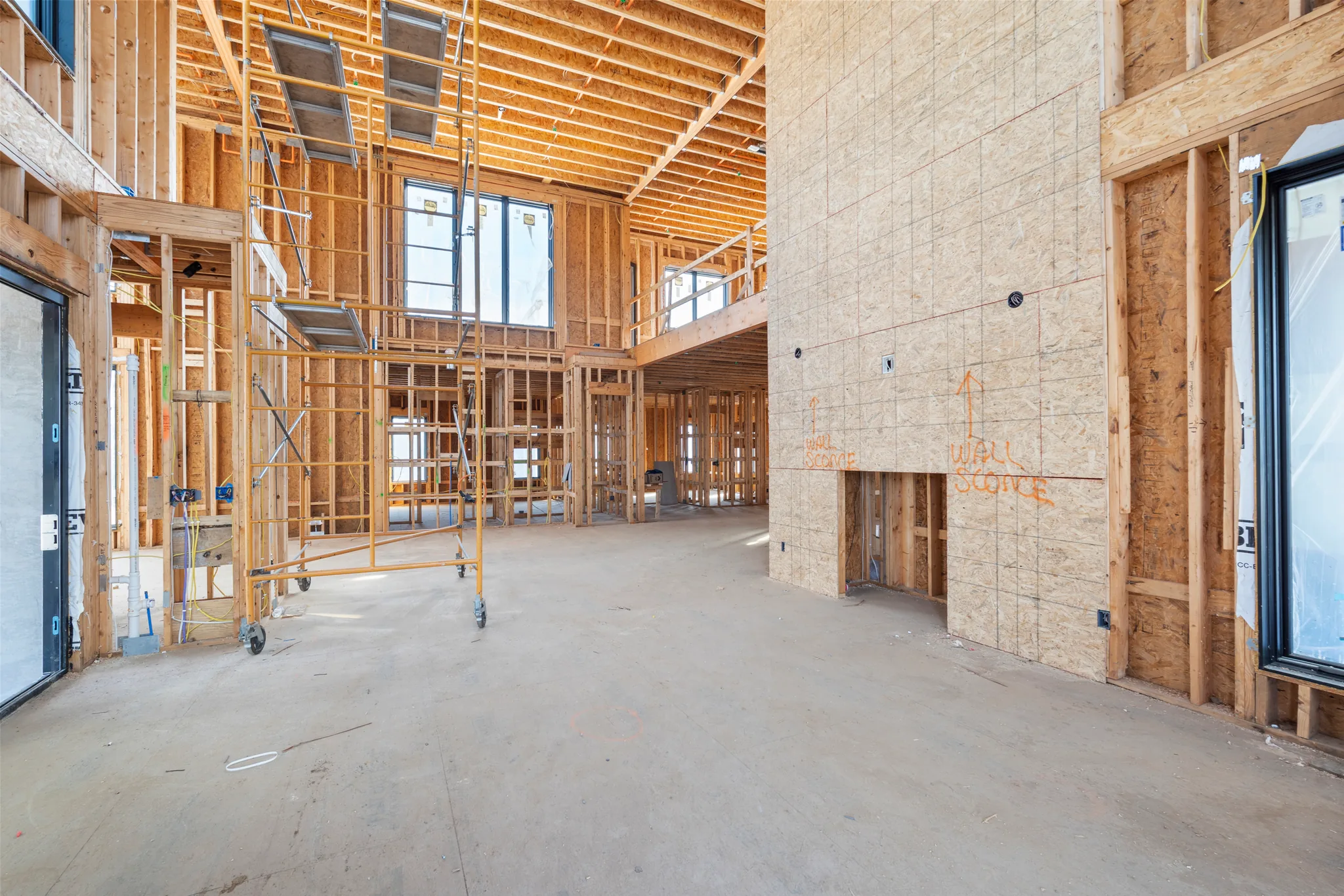 Two-story interior framing under construction with an OSB-sheathed fireplace wall marked for wall sconces, rolling scaffold and tall black windows