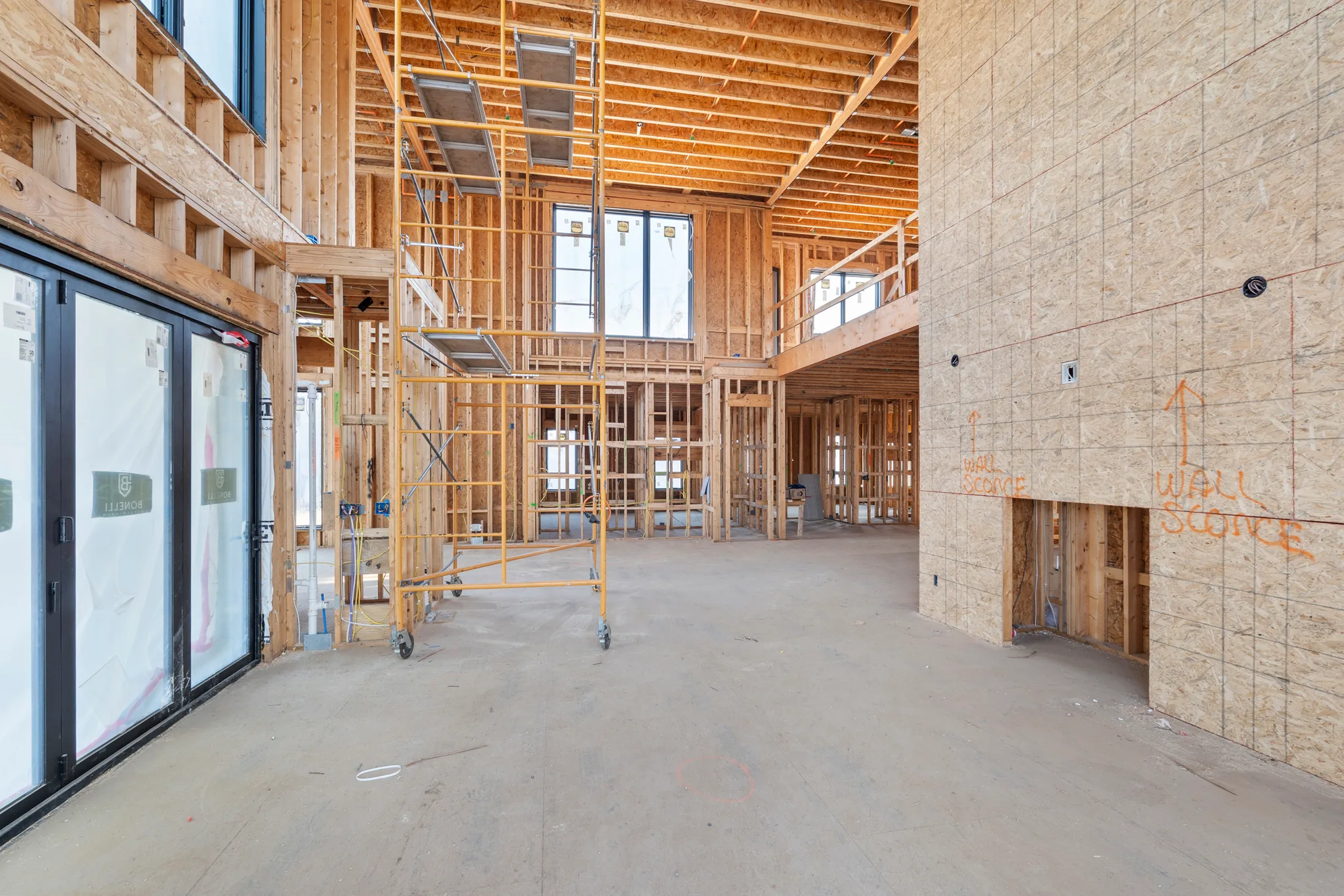 Interior framing under construction with a tall OSB-sheathed wall marked for wall sconces, rolling scaffold, black sliding doors and concrete floor