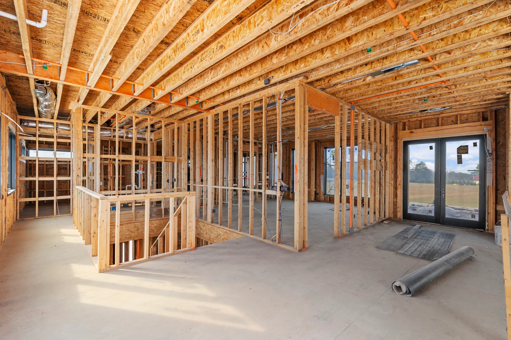 Interior of a home under construction with exposed wood stud walls, OSB floor joists above, concrete slab and installed black French doors