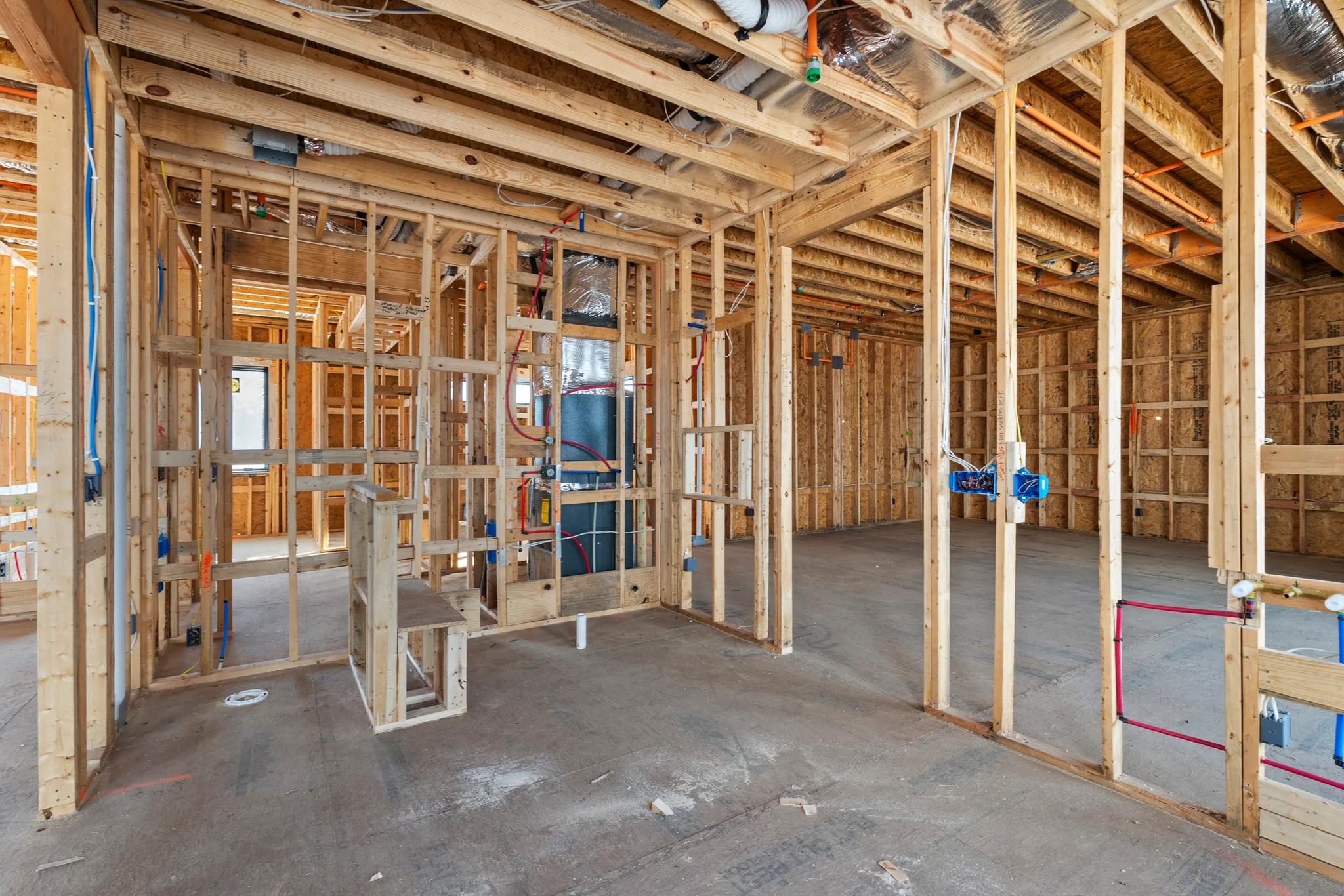 Interior wood framing under construction with rough-in wiring, red and blue water lines, ductwork and blue electrical boxes over a concrete slab