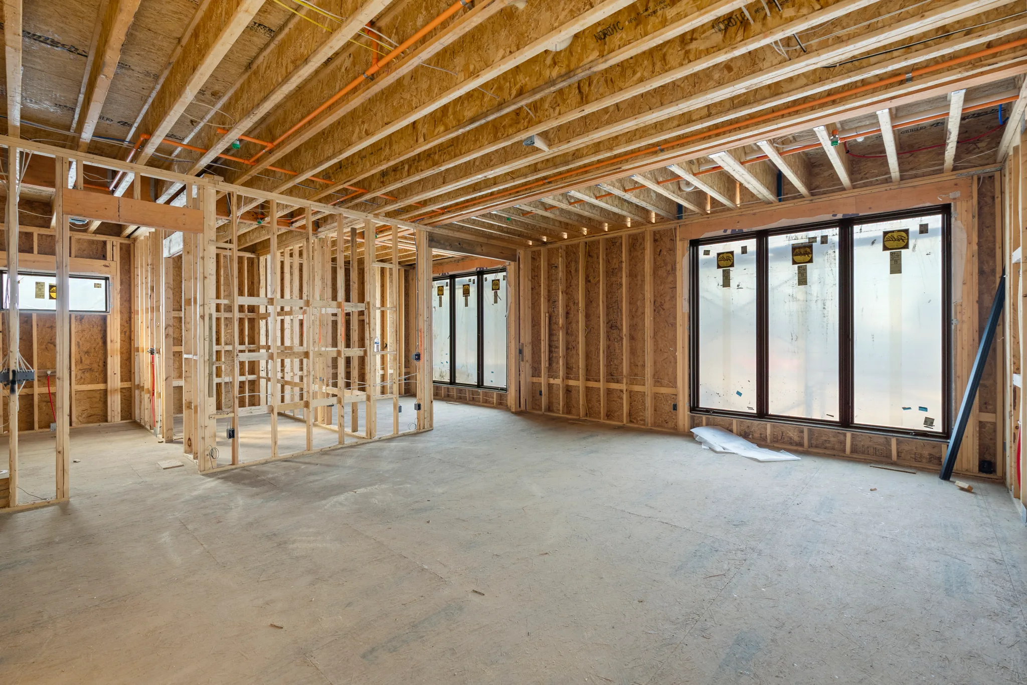 Interior under construction with exposed floor joists, stud partition walls, orange conduit overhead, and large black window units on a concrete floor