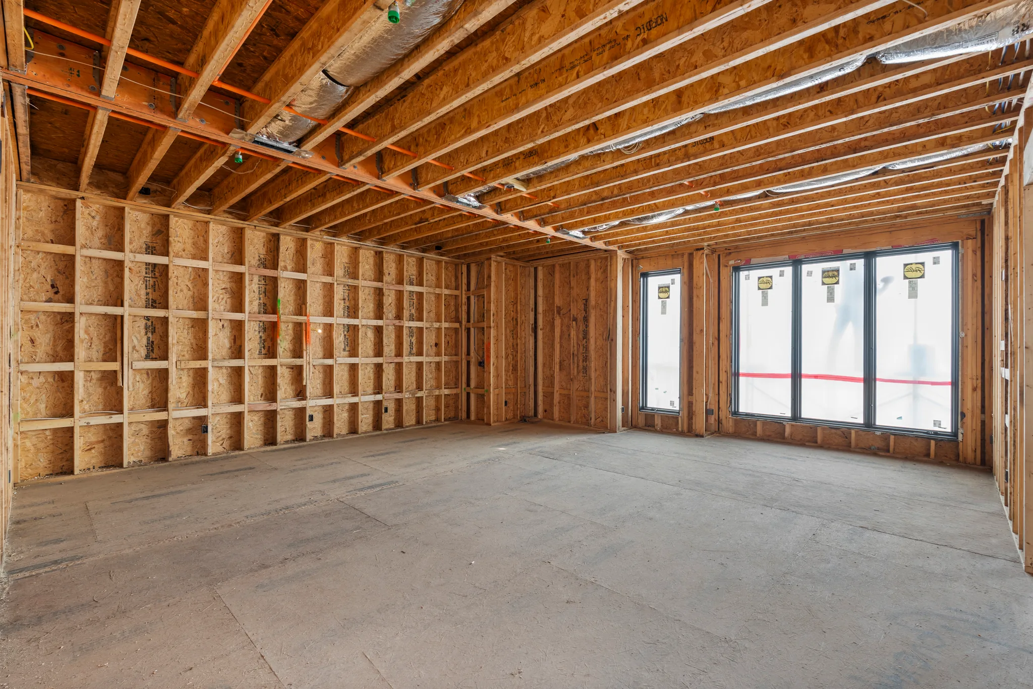 Interior room under construction with exposed wood stud walls, OSB sheathing, ceiling joists with insulated ducts and new black-framed windows above a concrete floor