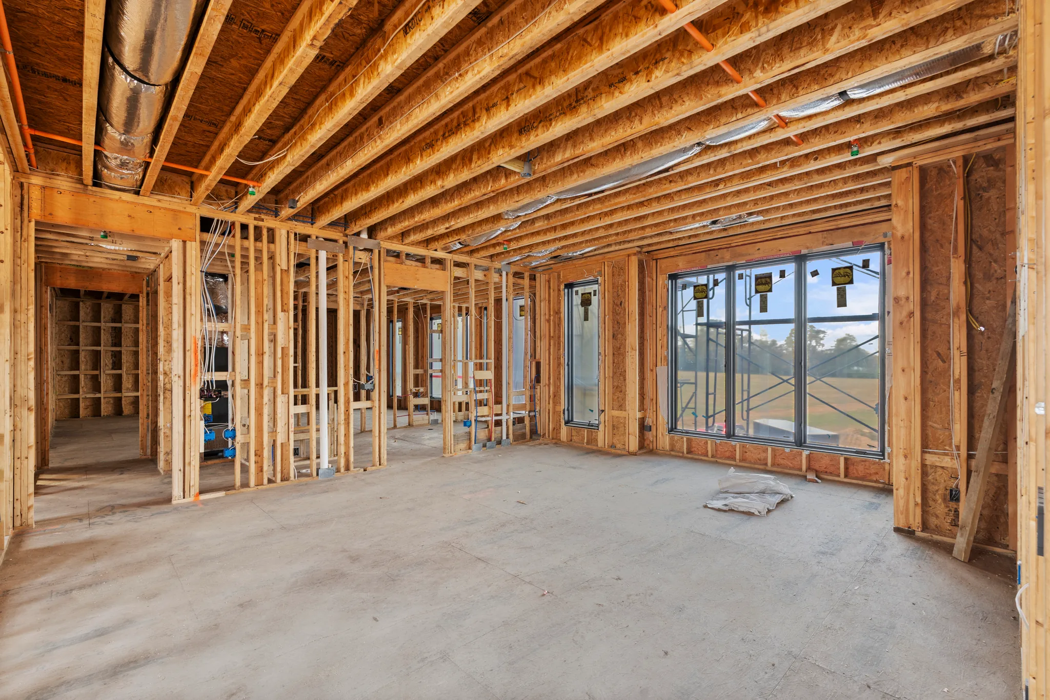 Interior wood framing under construction with exposed joists, ductwork, wiring and installed black windows above a bare concrete floor