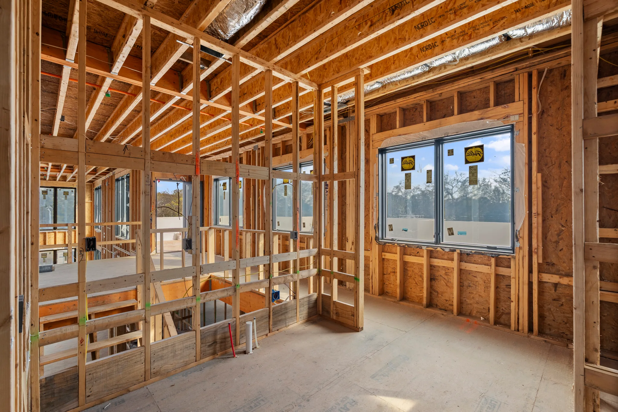 Interior wood framing under construction with black-framed windows installed, exposed joists overhead, insulated duct runs and bare subfloor lit by daylight