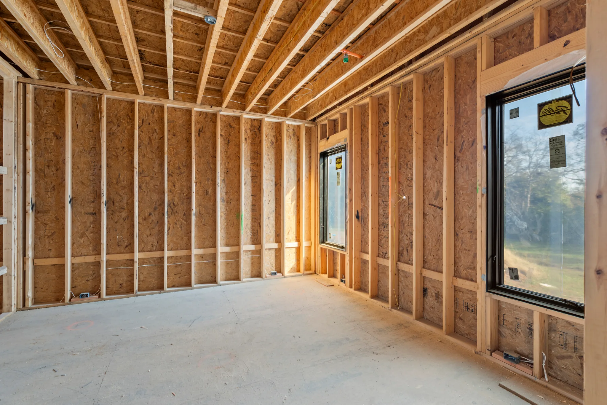 Interior room under construction with wood stud walls, OSB sheathing, exposed ceiling joists and new black windows still carrying manufacturer labels