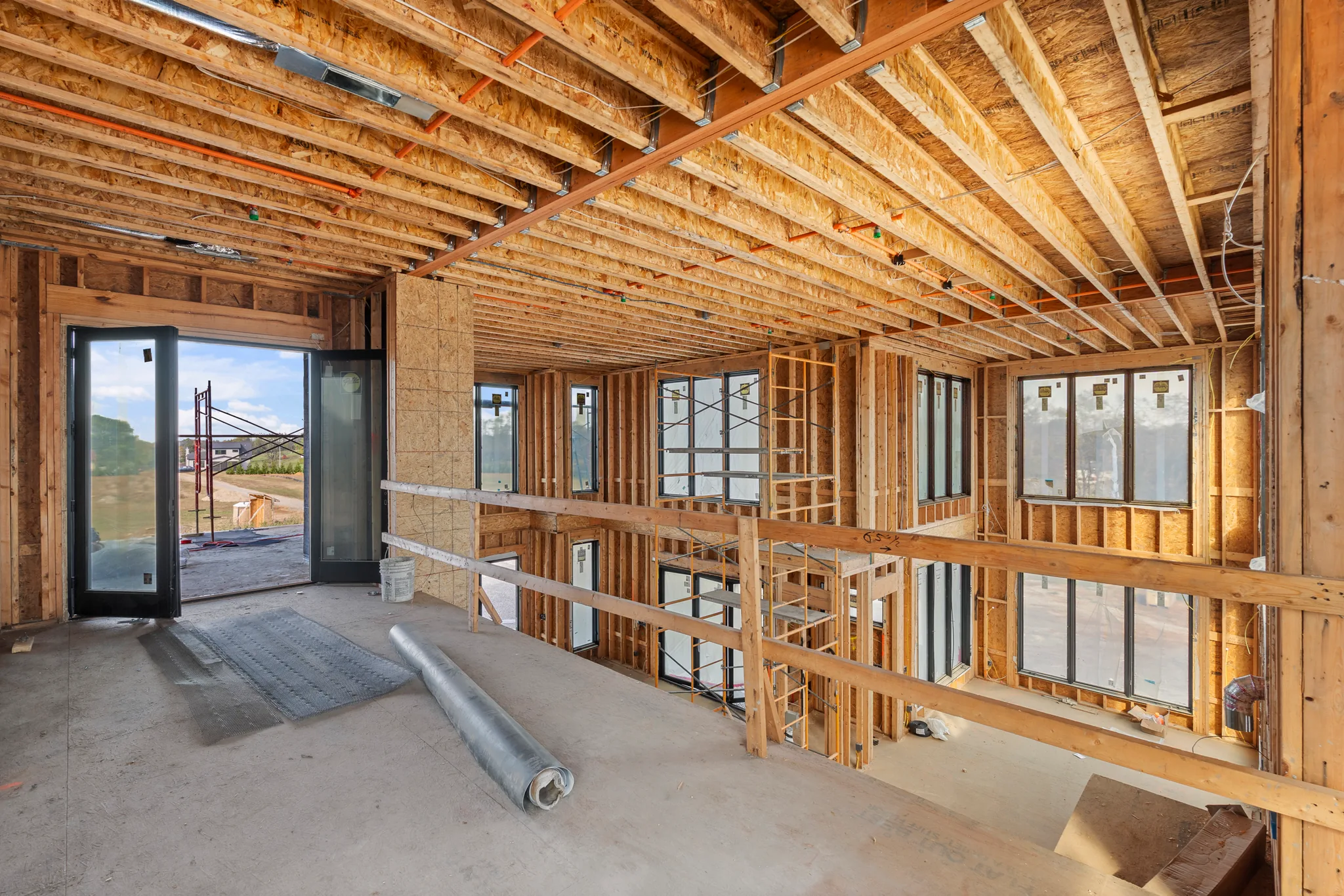 Interior framing under construction with exposed joists, sheathing and black-framed windows, with open glass doors leading to a graded site