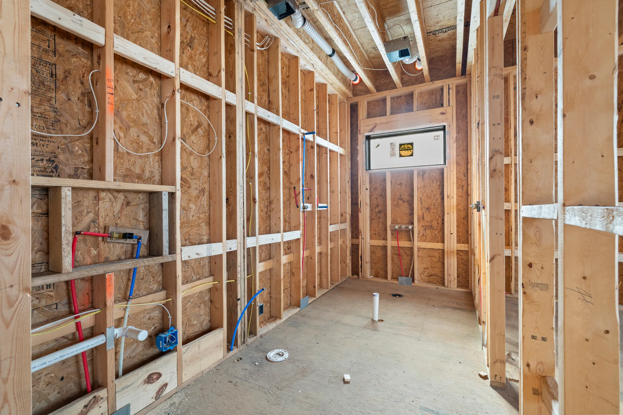 Narrow framed room under construction with OSB sheathing, red and blue PEX supply lines, wiring, and a single window opening