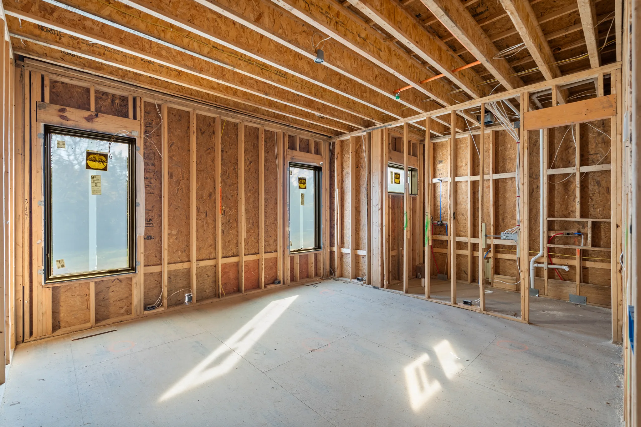 Interior room under construction with stud walls, installed windows, exposed ceiling joists and plumbing and wiring rough-ins on a concrete slab
