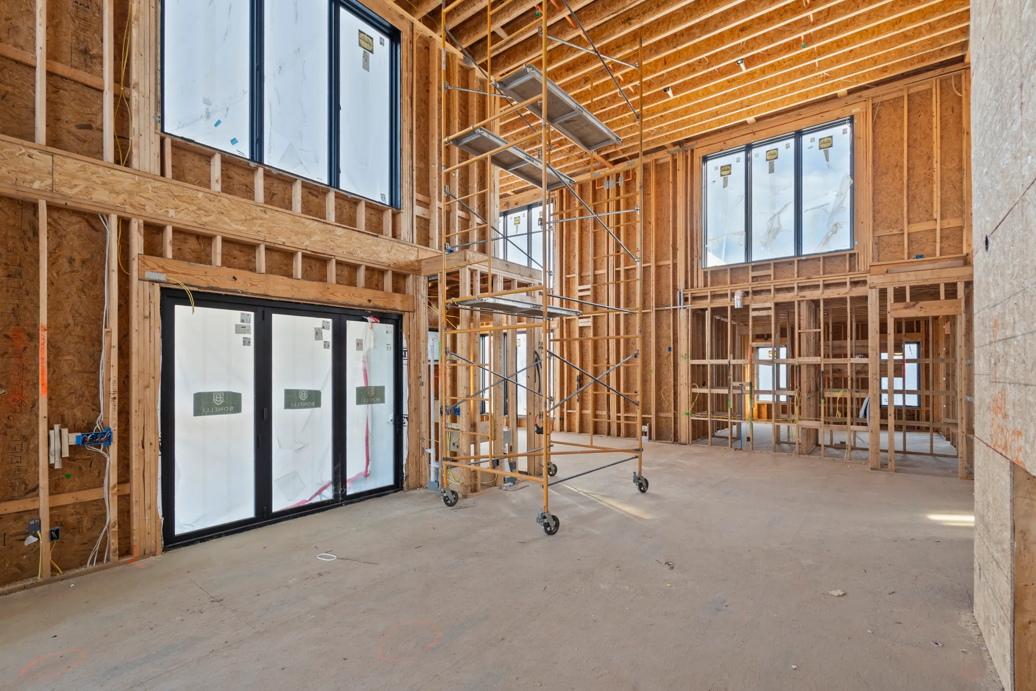 Interior framing under construction with black-framed windows and sliding doors still covered in protective film, rolling scaffolding and a concrete floor