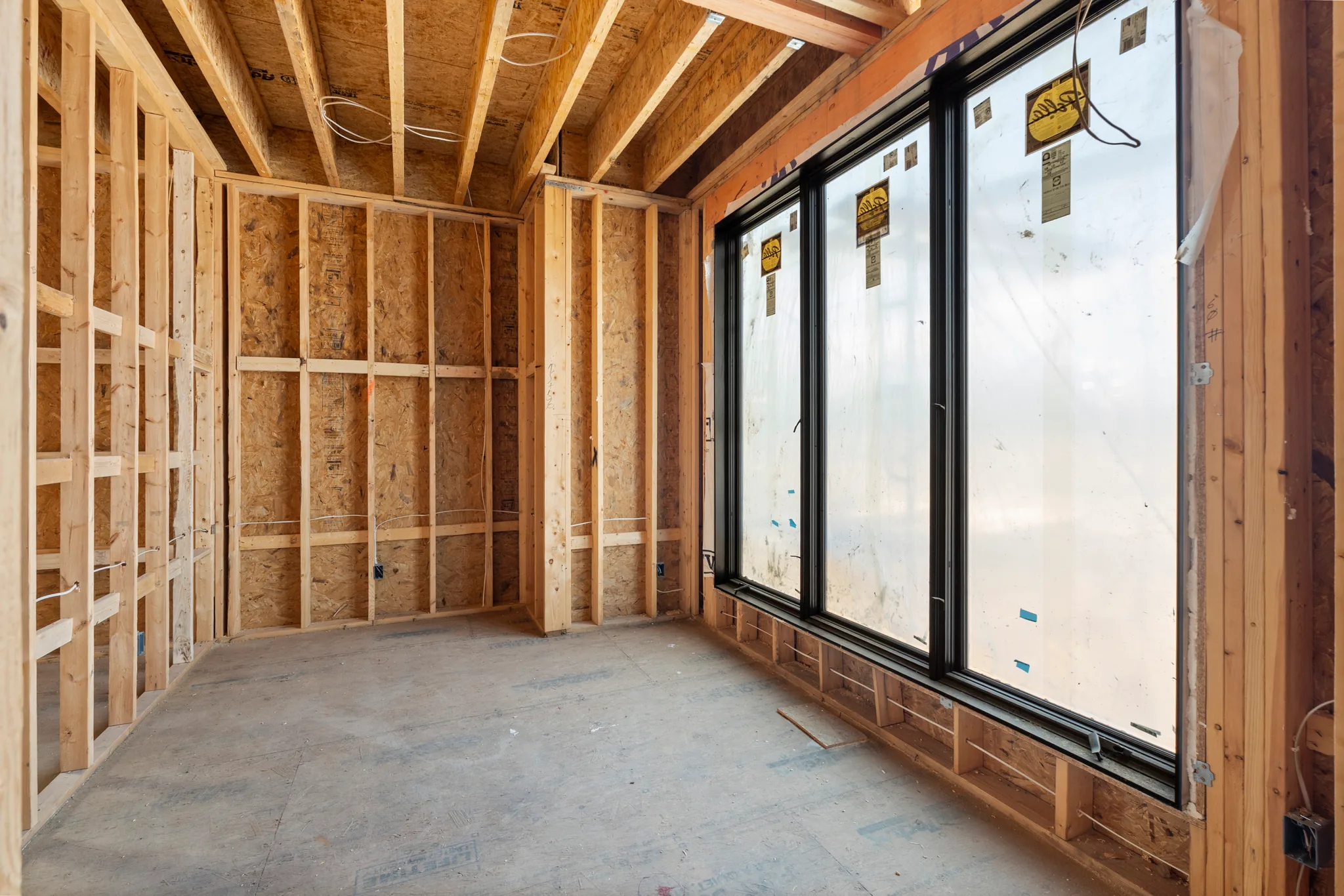 Framed interior room under construction with OSB sheathed walls, exposed ceiling joists and a wall of tall black-framed windows still labeled