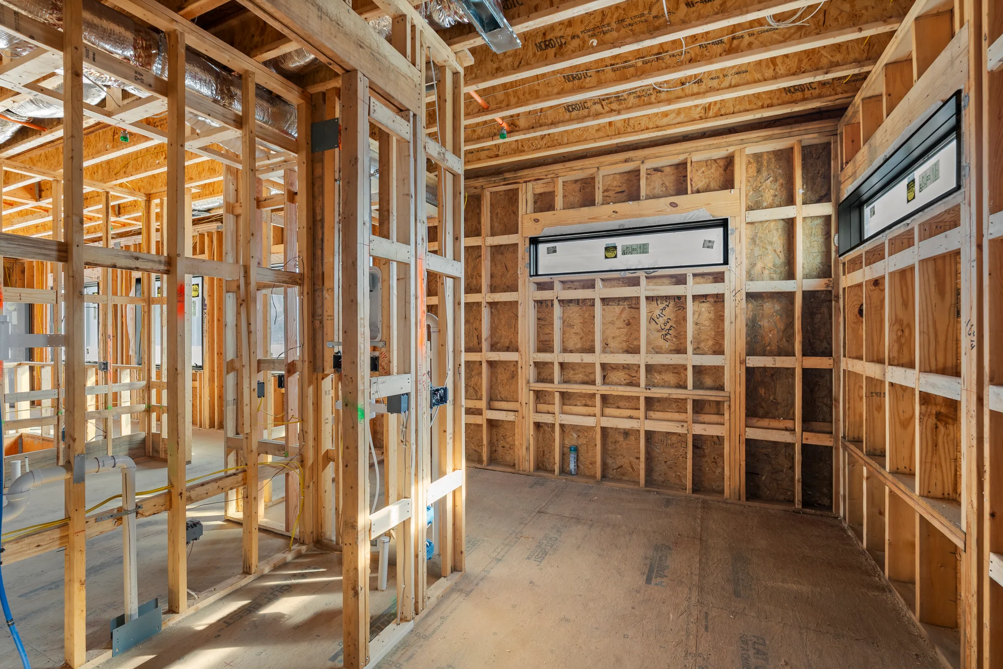 Interior wood framing under construction with plywood sheathing walls, two wrapped black window units, exposed ductwork and rough wiring on a plywood floor