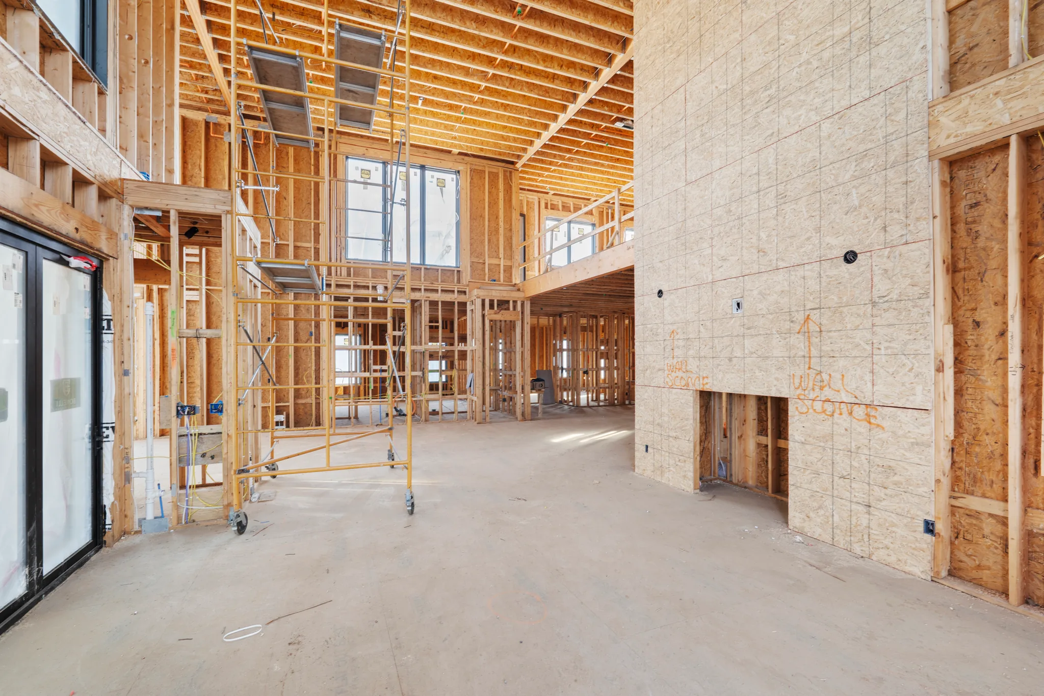 Interior of a home under construction with two-story framed great room, OSB sheathed fireplace wall marked wall sconce, rolling scaffold and concrete floor
