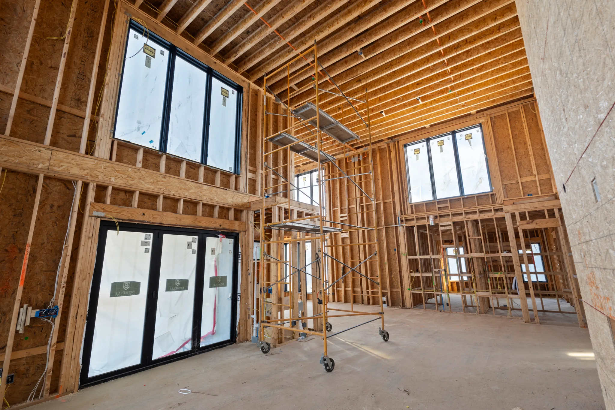 Interior of a home under construction with exposed stud walls, open ceiling joists, black windows still labeled, and a rolling scaffold on a concrete slab