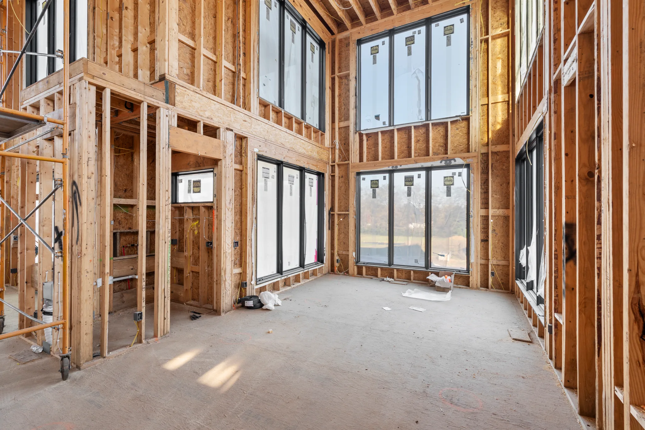 Interior framing under construction with stacked black window units on two levels, plywood subfloor, scaffolding at left and views of open land outside