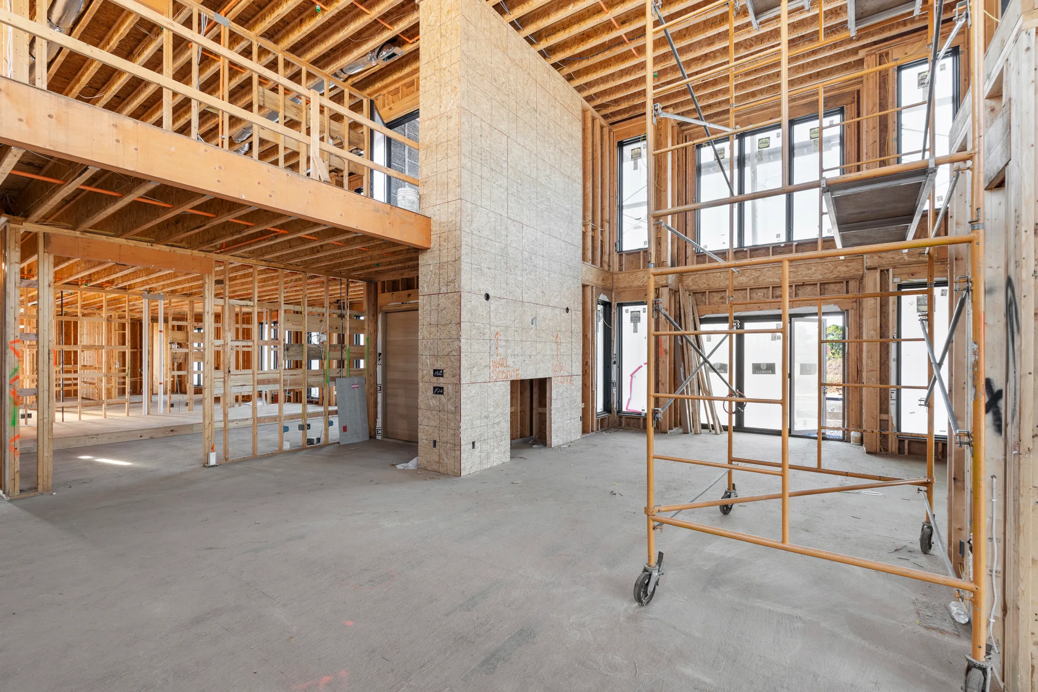 Interior of a home under construction with two-story framing, a sheathed chimney chase, black window units and rolling scaffold on a concrete floor