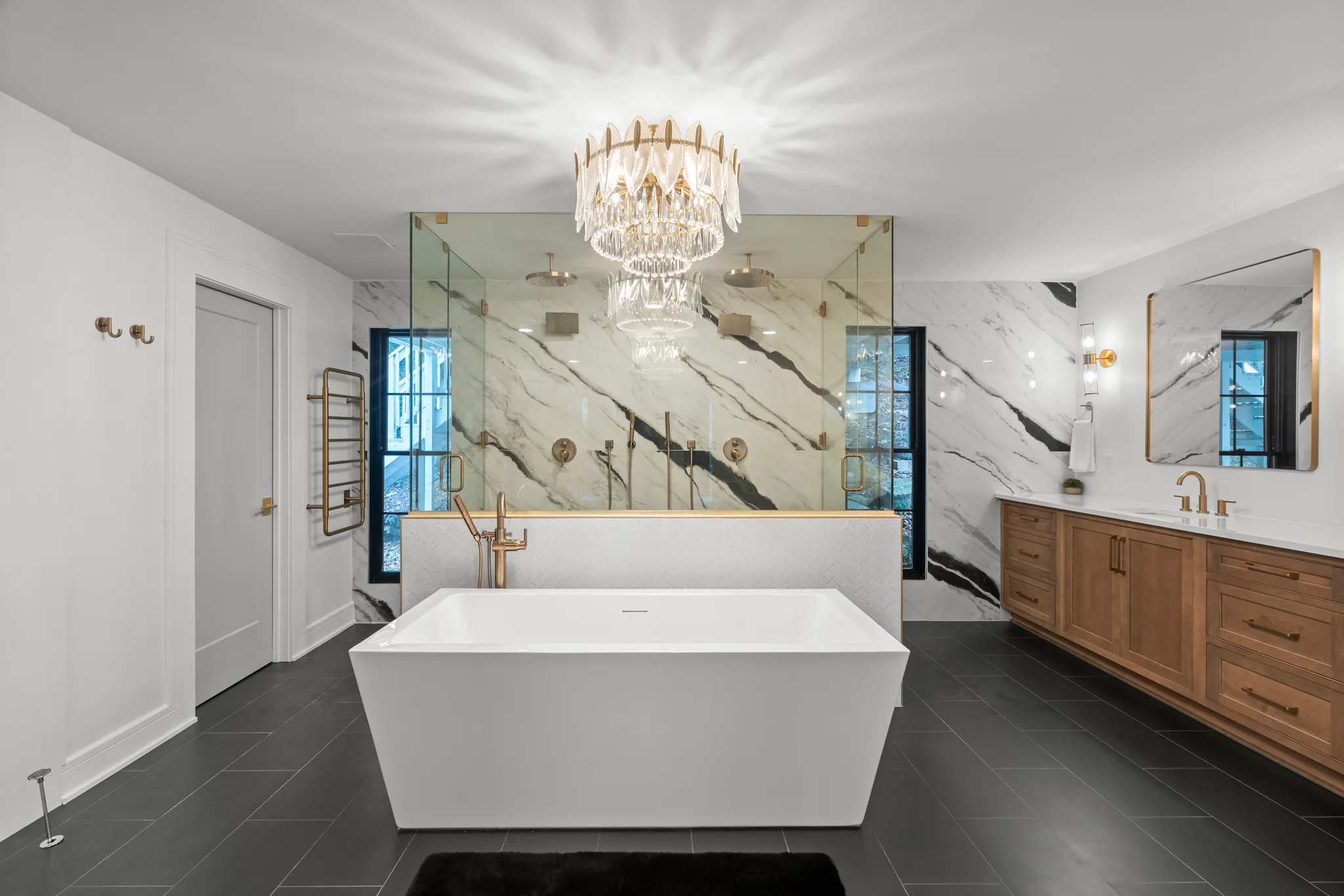 Primary bath with rectangular freestanding tub, glass shower behind veined marble walls, wood double vanity and dark gray floor tile