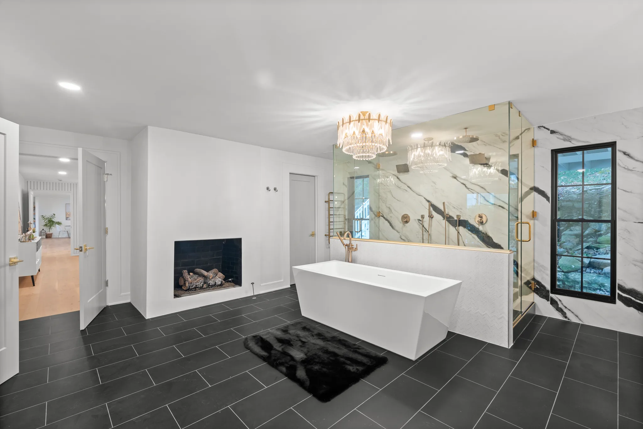 Primary bath with a freestanding rectangular tub, black-tiled fireplace, tiered crystal chandelier, marble-look shower wall and dark gray slate floor tile