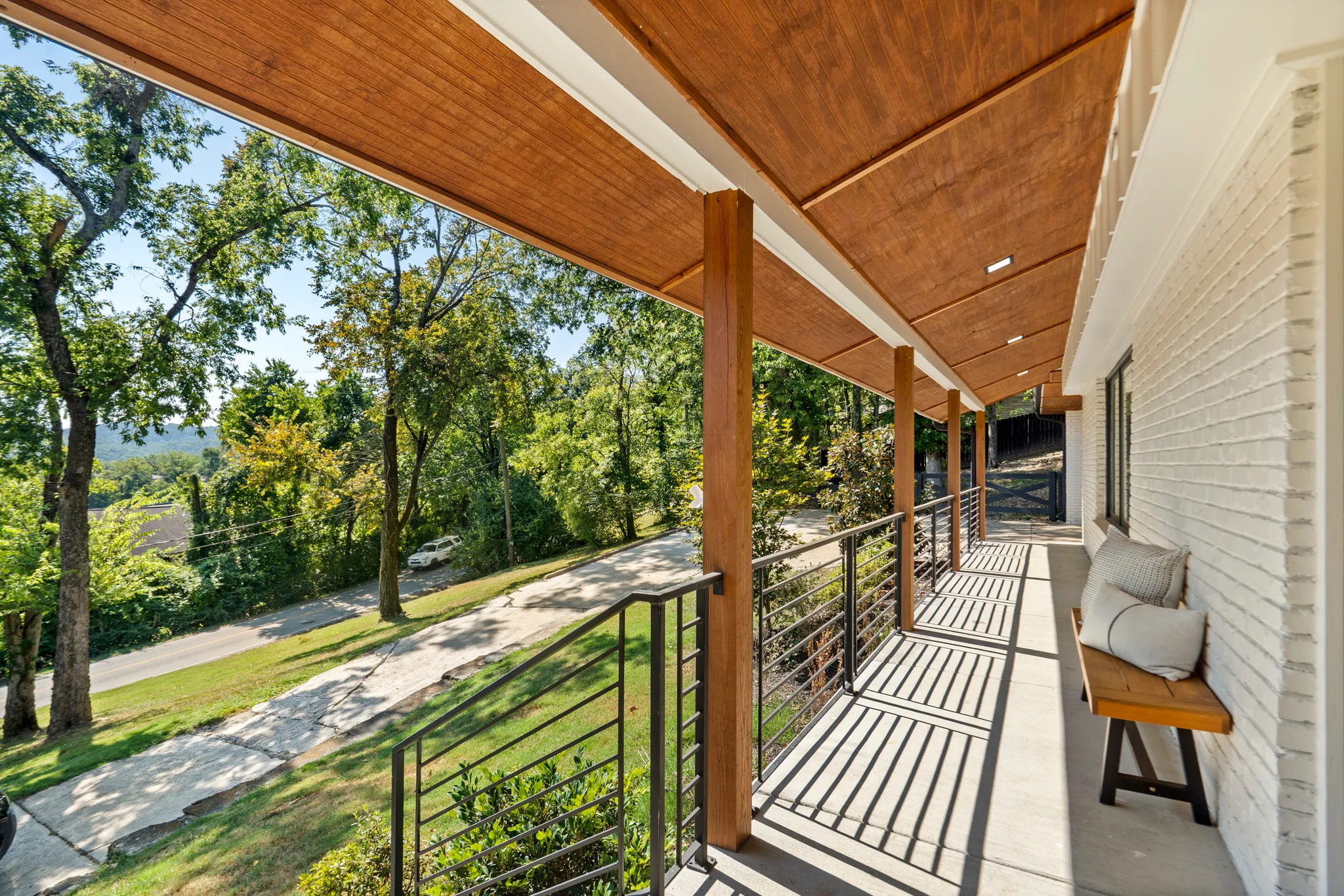 Long covered front porch with stained wood ceiling, timber posts, black horizontal metal railing, painted white brick wall, and a wood bench