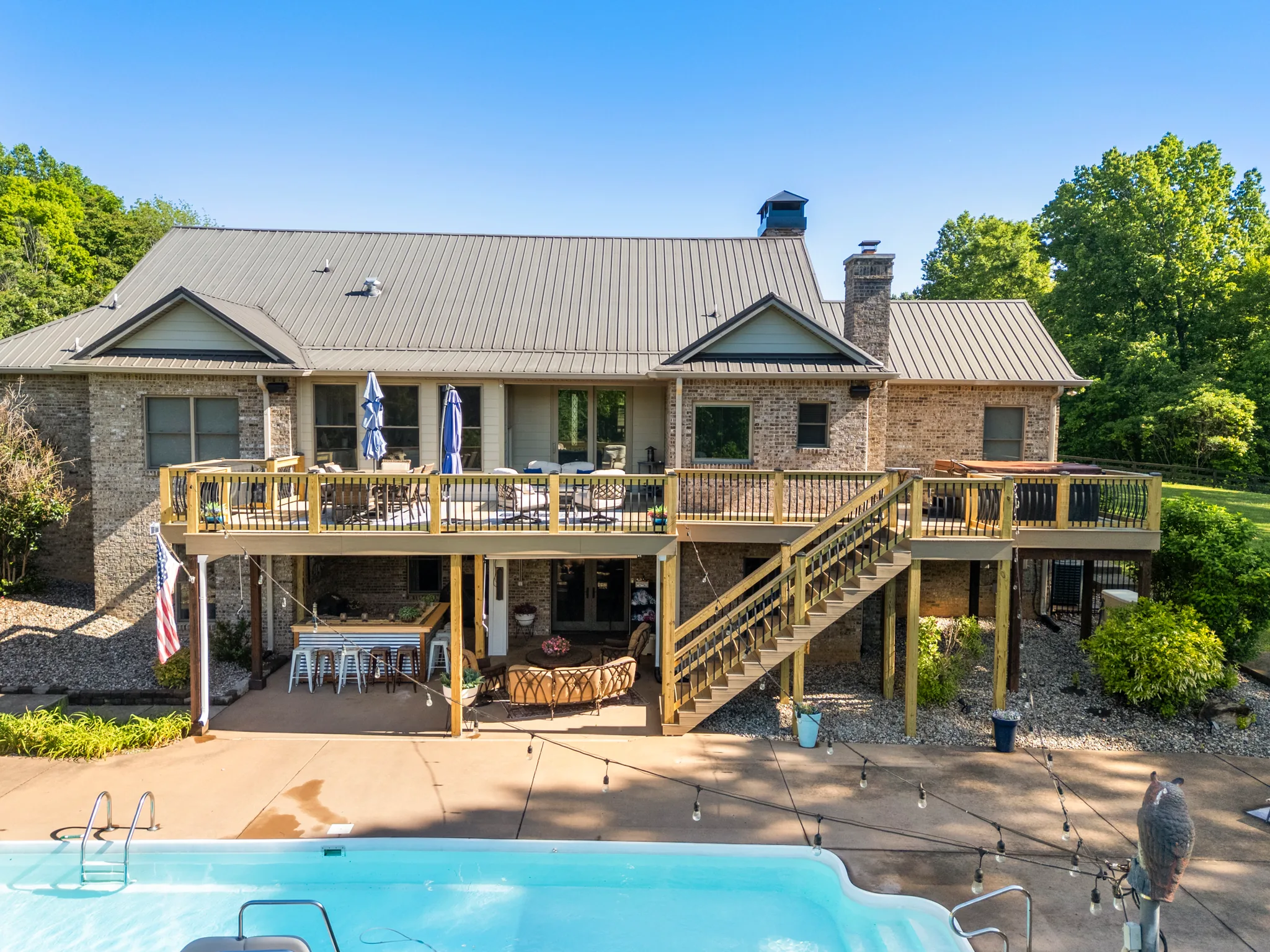 Wood deck spanning the rear of a brick and board-and-batten home with metal roof, stairs down to a concrete pool patio