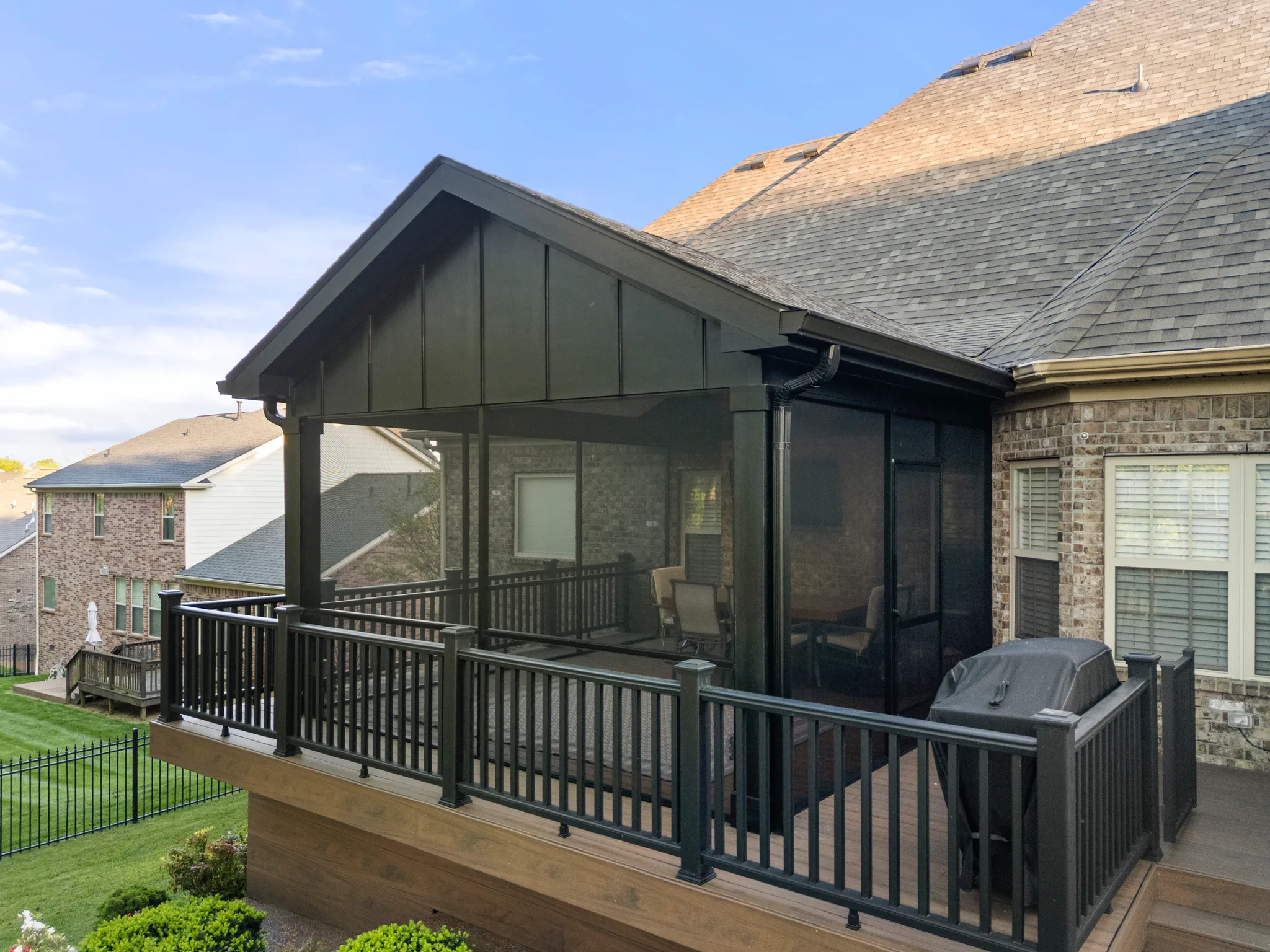 Screened porch with dark gable roof and board and batten gable end attached to a brick home, next to a composite deck with black railings