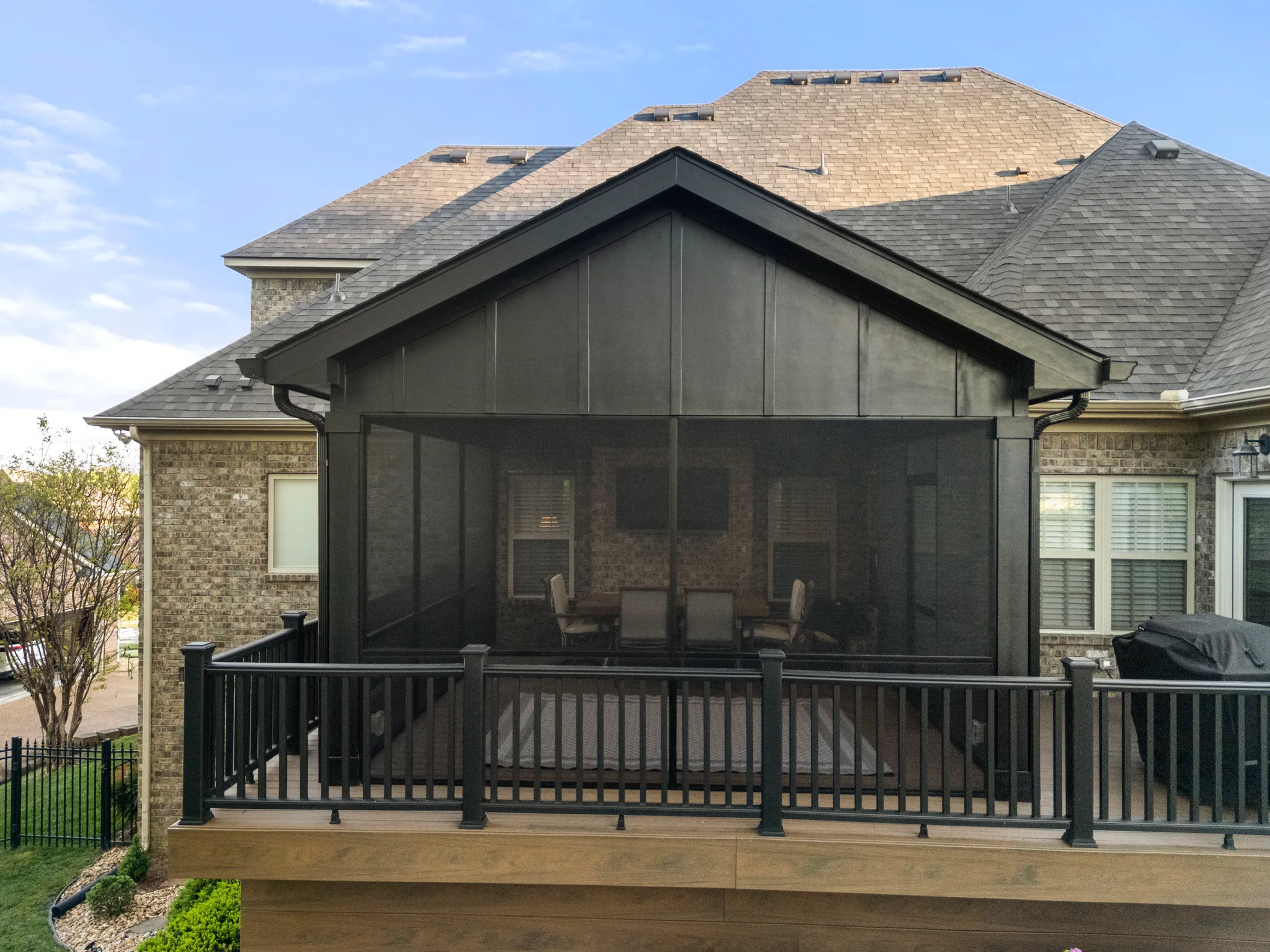 Screened porch with a dark gable roof and black screening attached to a brick home, fronted by a composite deck with black metal railing