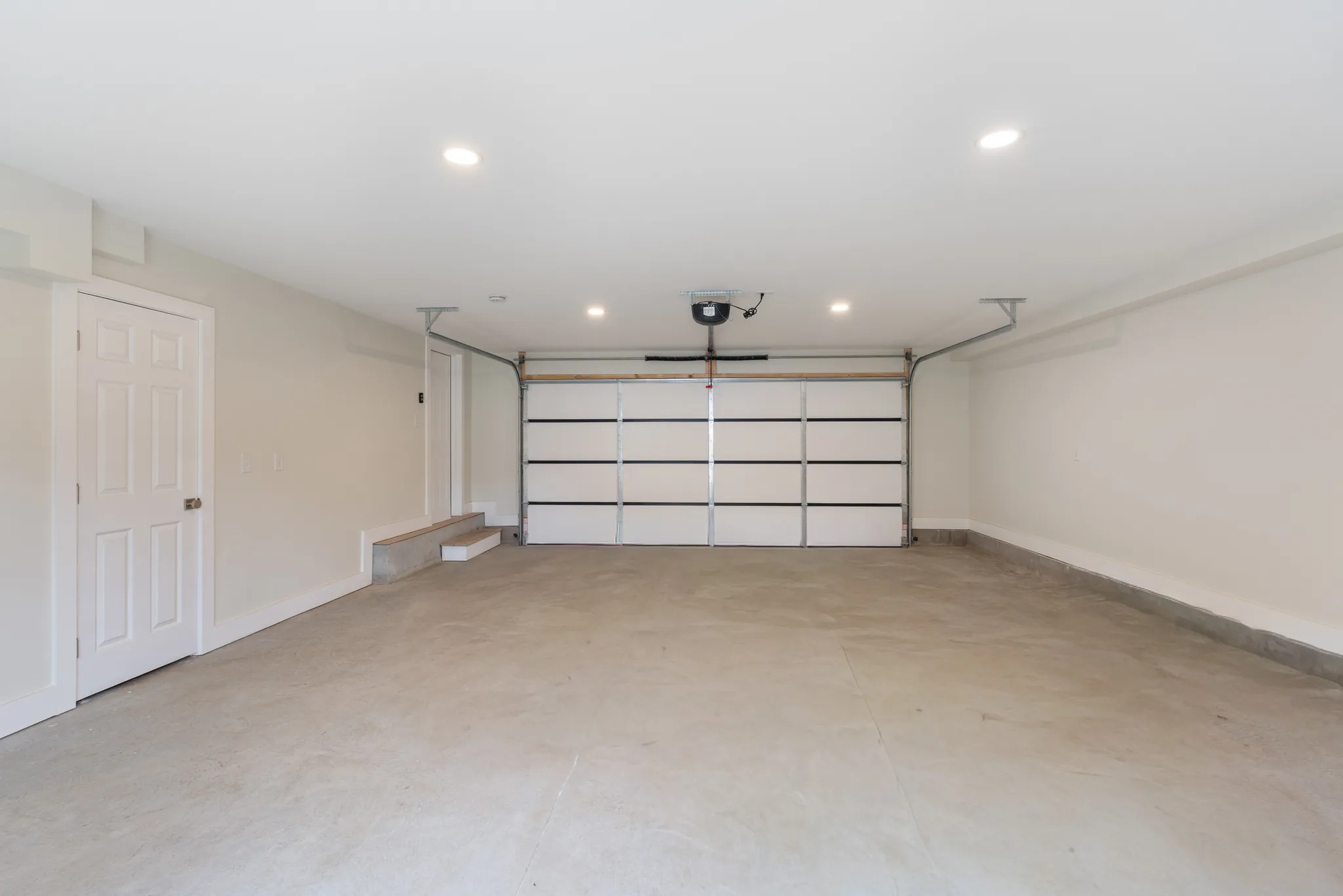 Finished garage interior with smooth concrete floor, cream walls, white sectional overhead door, recessed ceiling lights and a step up to an interior door