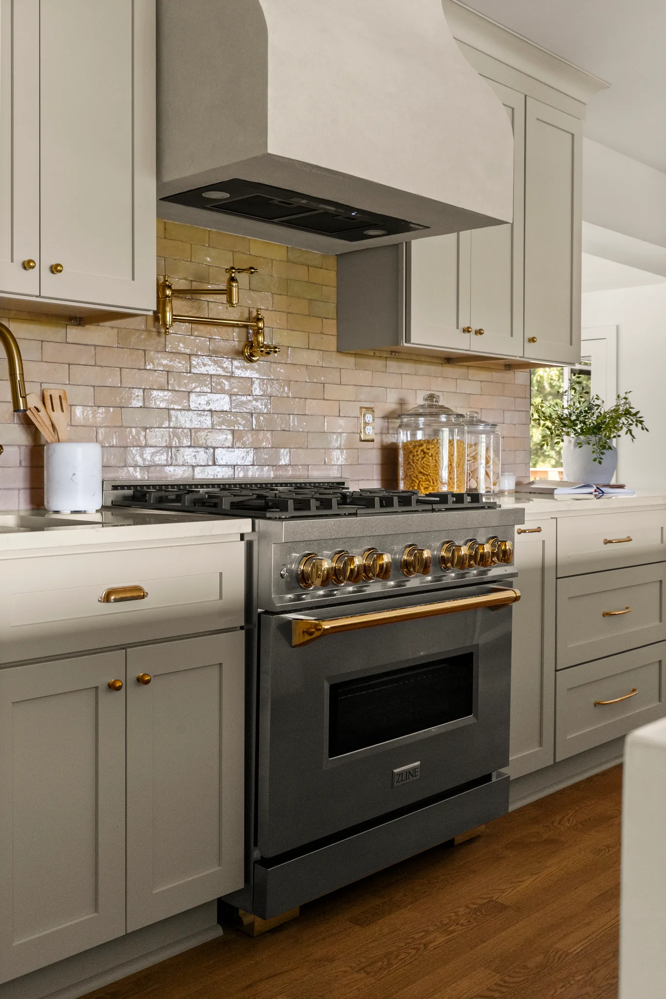 Kitchen range wall with a dark gas range trimmed in brass, glazed tile backsplash, greige shaker cabinets and oak floors
