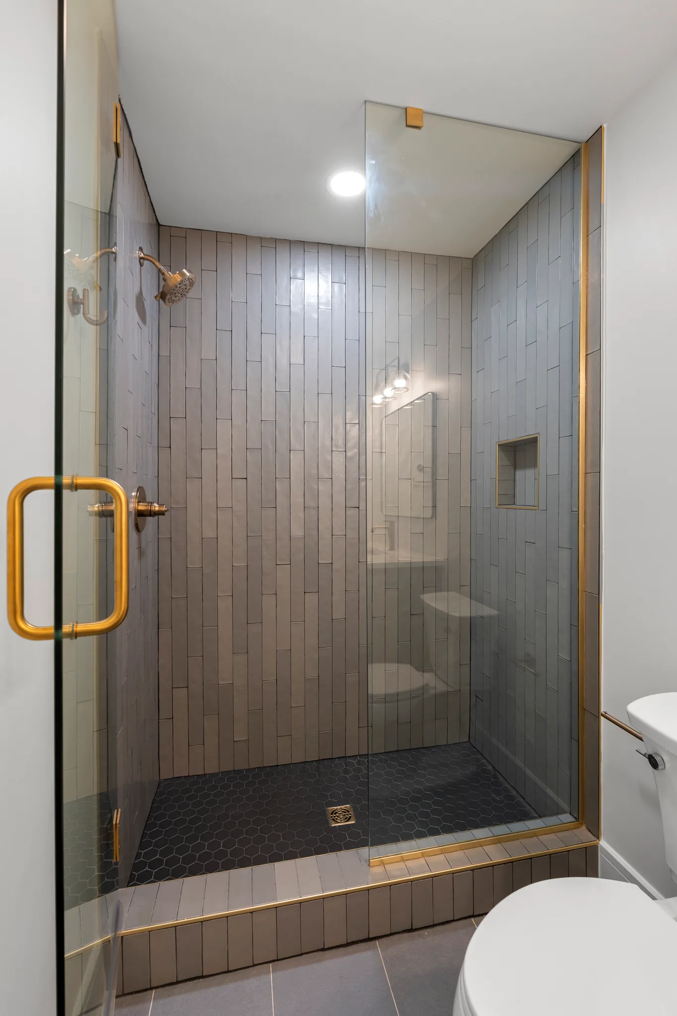 Walk-in shower enclosed in glass with vertical gray stacked tile, brass hardware, black hexagon floor tile and a recessed wall niche
