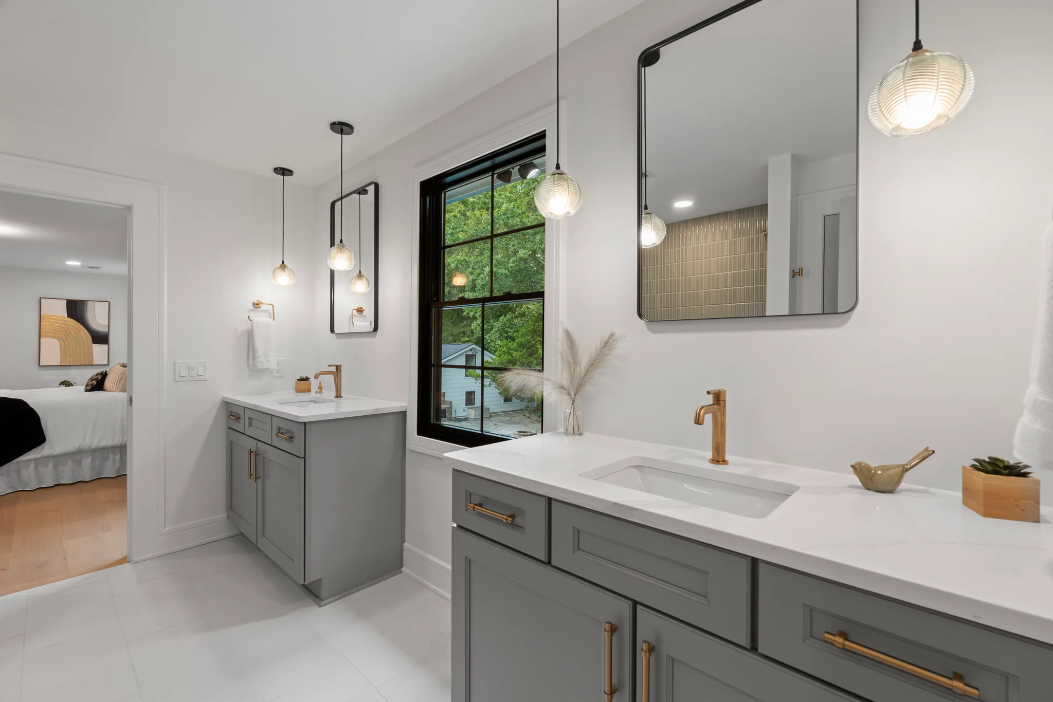 Primary bath with two gray shaker vanities, white quartz counters, brass faucets, glass pendant lights and a black-framed window