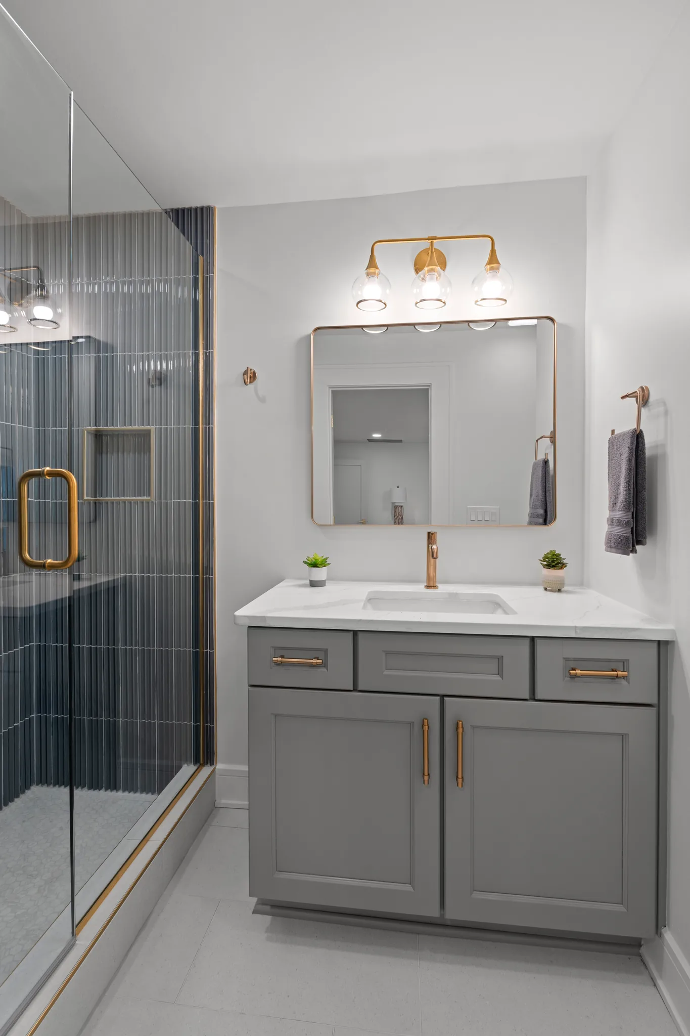 Bathroom with a gray shaker vanity, white quartz top, brass faucet and lighting, beside a glass shower lined with blue fluted tile