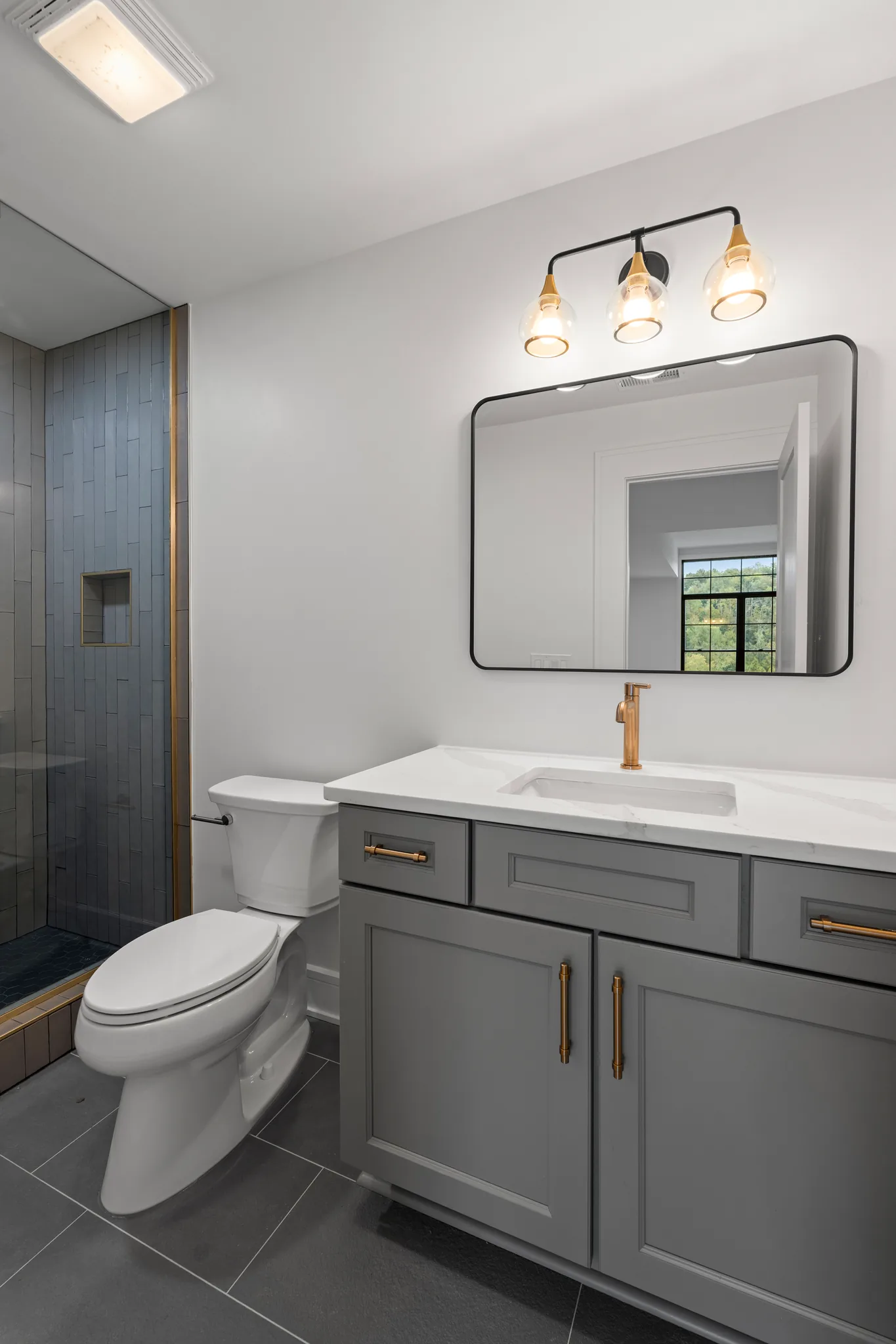 Bathroom with a gray shaker vanity, white quartz top, brass faucet, black-framed mirror, and walk-in shower lined with narrow gray tile
