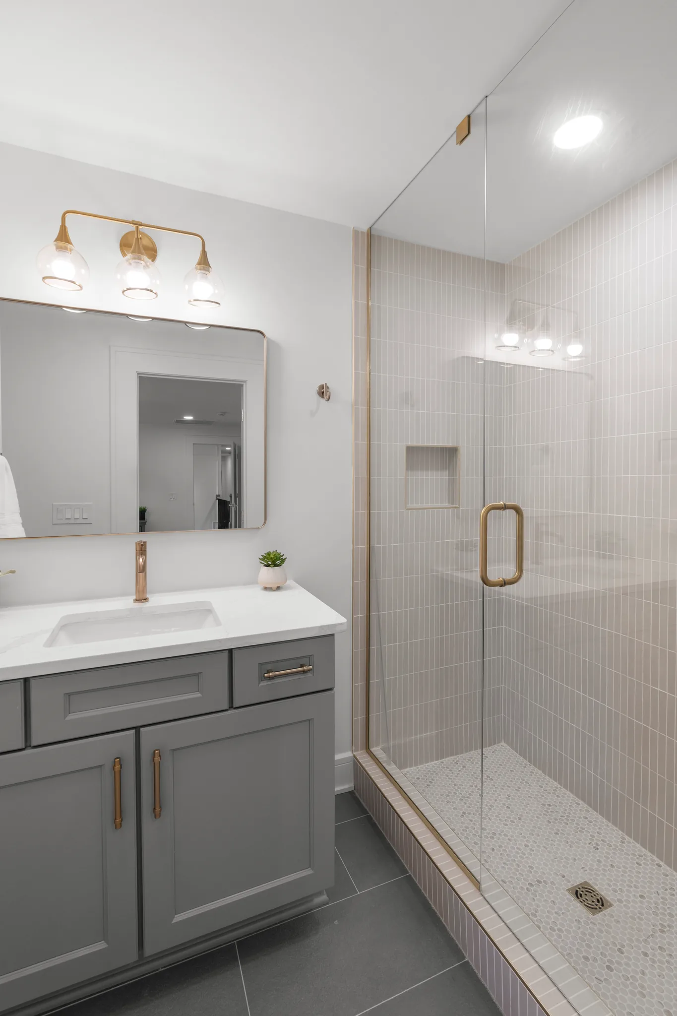 Bathroom with a gray shaker vanity, white quartz top, brass faucet and lighting, next to a glass shower lined in cream stacked tile