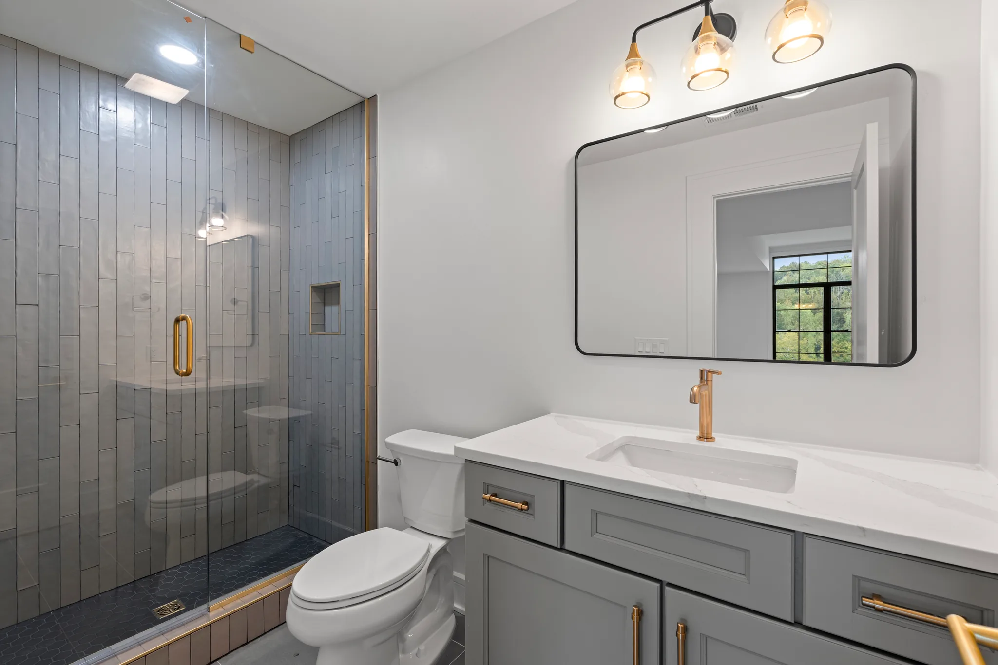 Bathroom with gray shaker vanity, white quartz top, brass faucet, black-framed mirror and glass shower lined with vertical gray tile